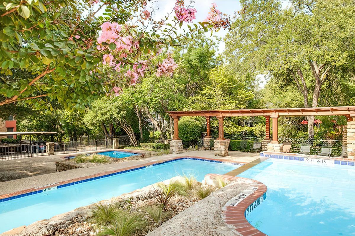 A serene outdoor pool area surrounded by lush greenery and blooming flowers, featuring two swimming pools and a pergola with stone accents. The setting invites relaxation in a tranquil environment, perfect for enjoying sunny days.