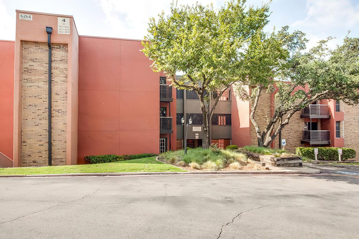 Exterior view of a modern apartment building with brick and stucco facades. The building features multiple floors, balconies, and lush greenery, including trees and shrubs, in the surrounding area. The driveway shows a smooth surface, leading to the entrance of the complex.