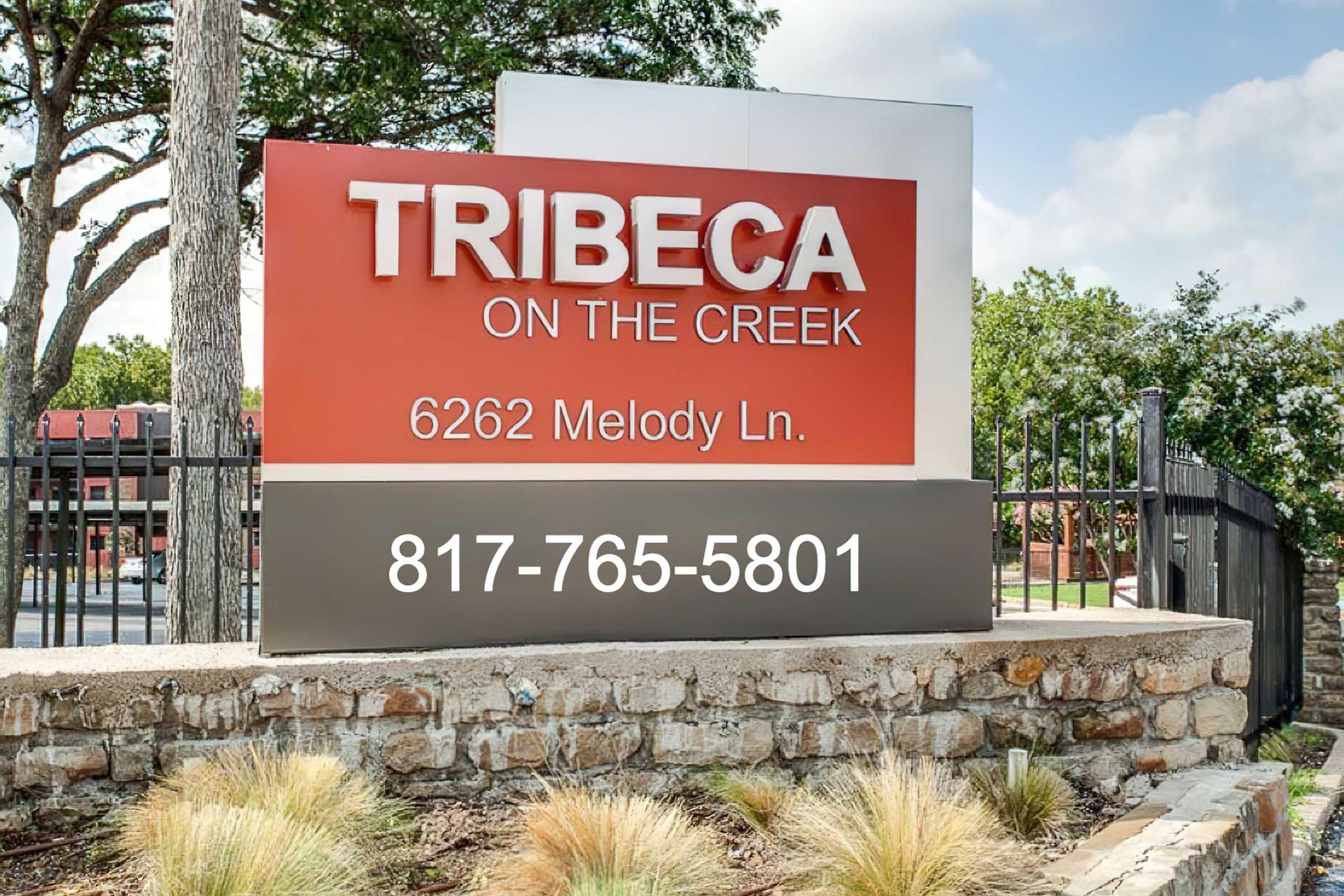 Sign for Tribeca on the Creek at 6262 Melody Ln, featuring a vibrant red background with white text displaying the name and phone number 817-765-5801. The sign is set against a backdrop of greenery and clear blue sky.