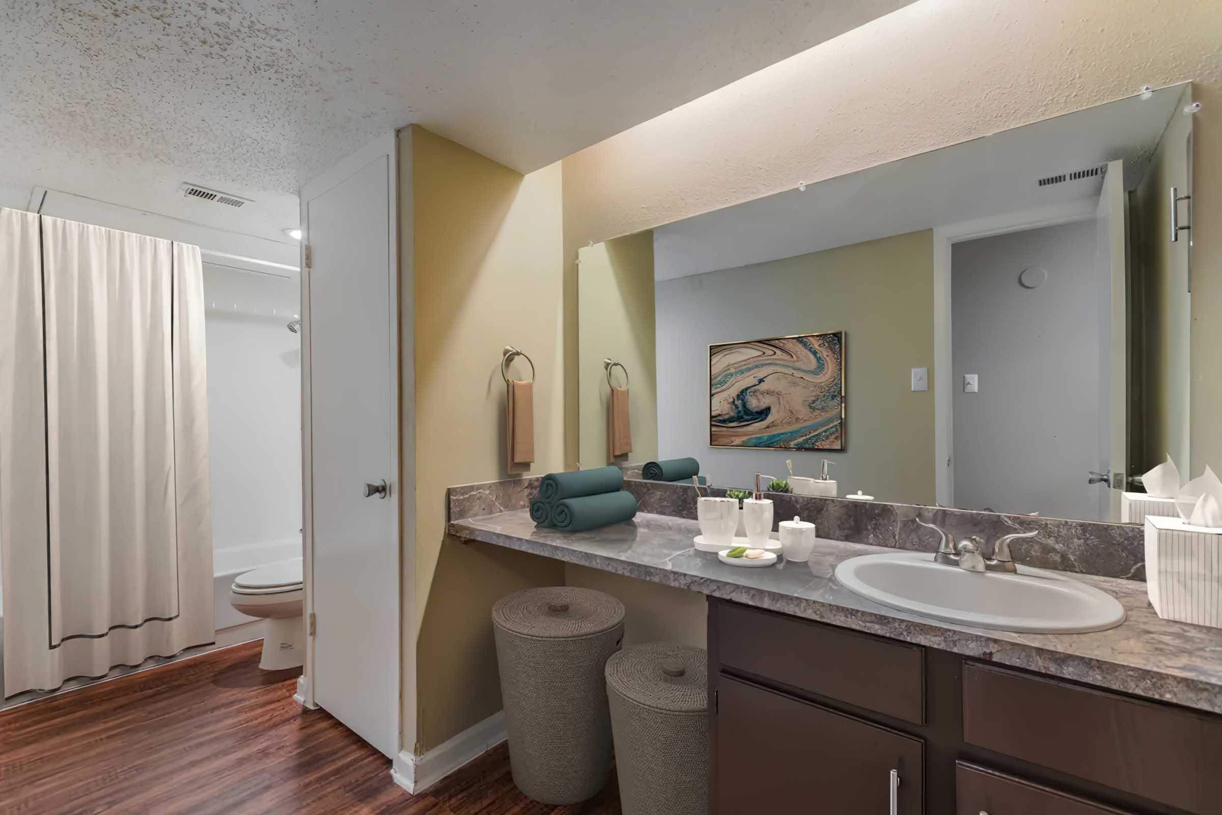 A modern bathroom featuring a large mirror above a sink with toiletries, rolled towels, and decorative items. The space includes a shower curtain, a toilet, and two round storage bins. The walls are painted in a soft yellow color, and the flooring is a dark wood finish.