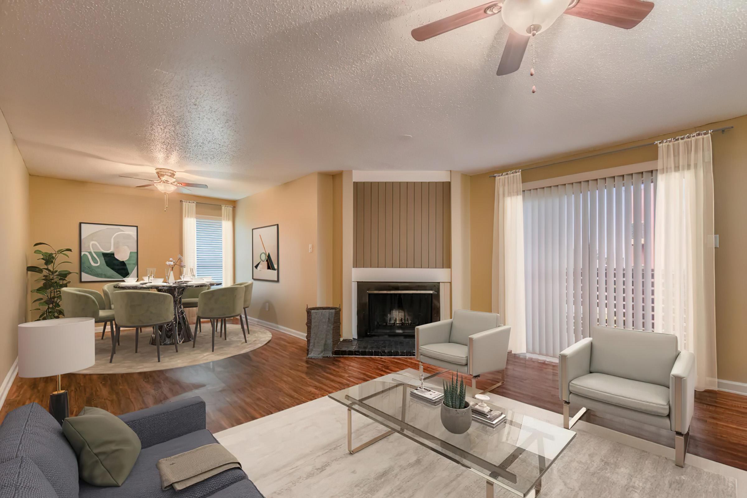 A bright and inviting living area featuring a comfortable blue sofa, a glass coffee table, and two light-colored chairs. A dining space with a round table and green chairs is visible in the background. Large windows allow natural light, and a cozy fireplace adds warmth to the atmosphere.