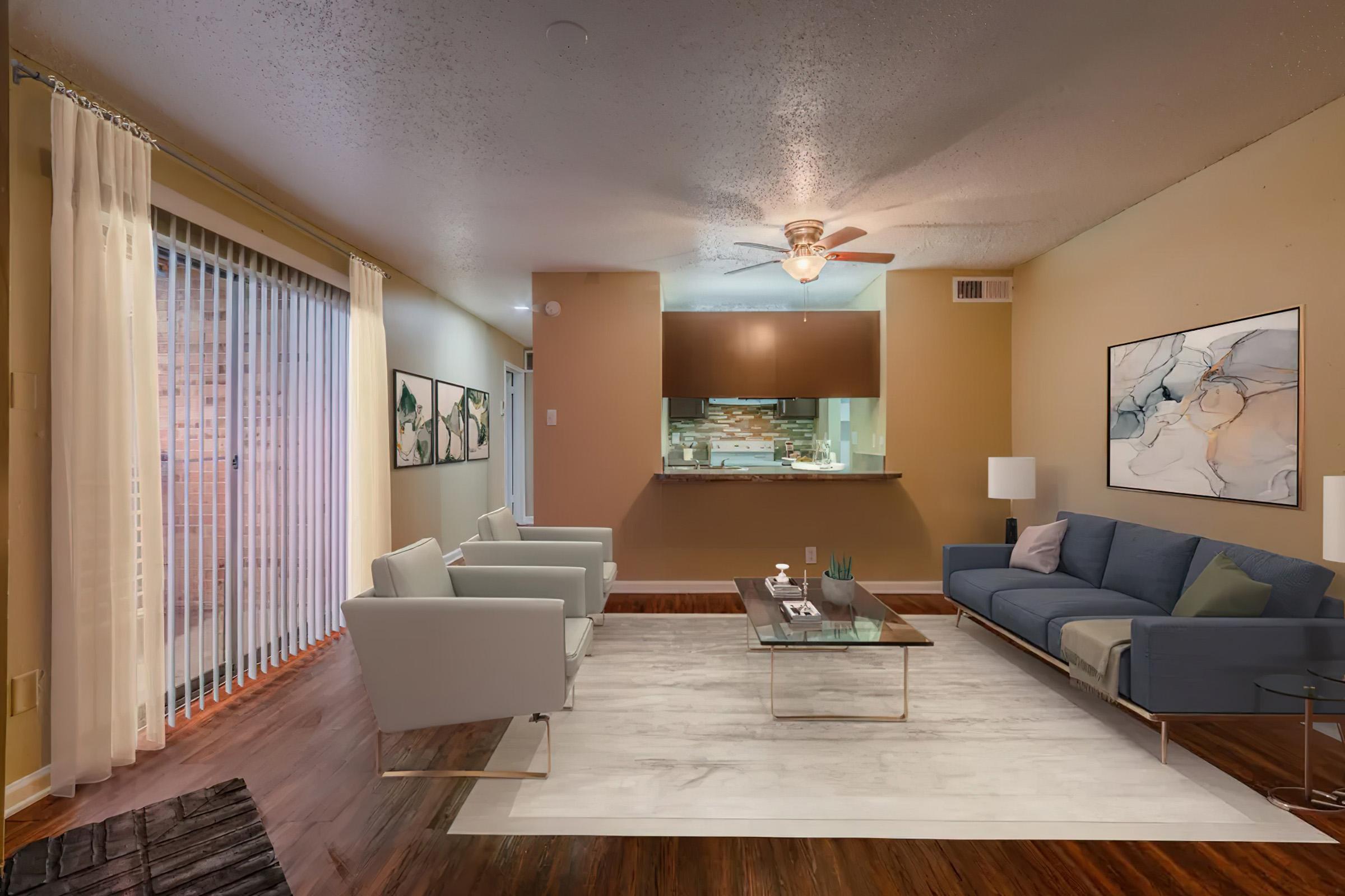 A cozy living room featuring a blue sofa, two light gray chairs, and a glass coffee table on a large white rug. The walls are painted in warm tones, with framed artwork displayed. A ceiling fan is visible, and behind the seating area, there is an open kitchen with a bar counter. Natural light comes through vertical blinds.