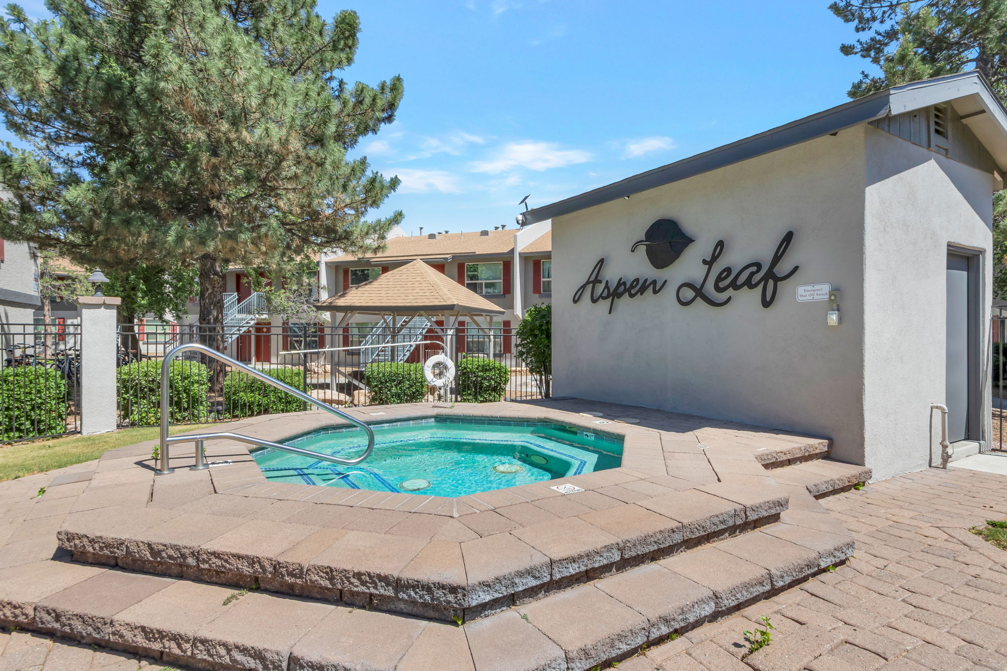 A small outdoor hot tub surrounded by stone steps, with a sign that reads "Aspen Leaf" nearby. Behind the hot tub, there are trees and a residential building, creating a relaxing atmosphere. The sky is clear and blue, adding to the inviting setting.