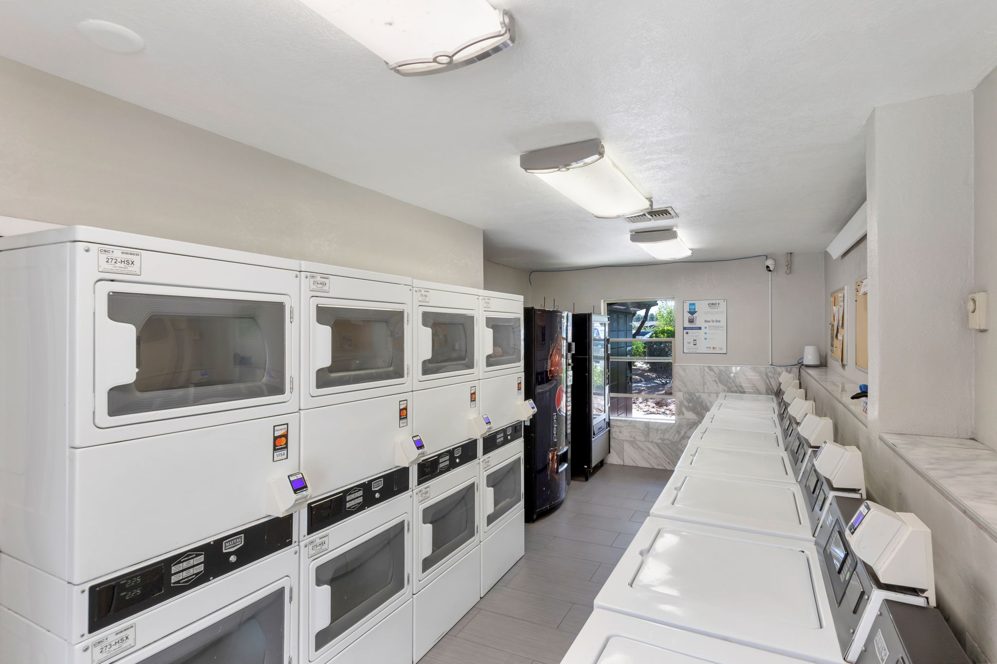 A clean, modern laundry room featuring several stacked washers and dryers along one wall, a vending machine to the side, and a large refrigerator. The space is well-lit with fluorescent lighting and has light-colored walls and flooring, creating a bright and functional environment for laundry.