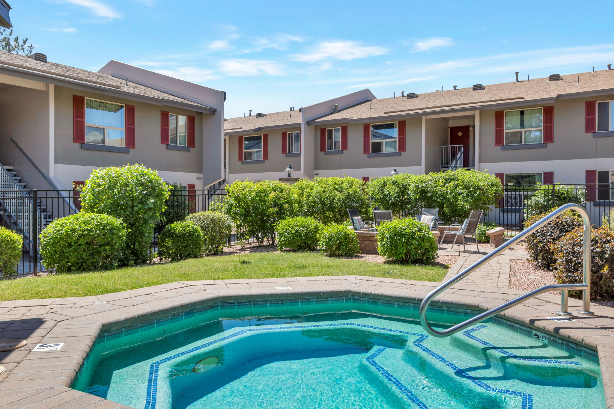 A view of an outdoor area featuring a small circular hot tub, surrounded by green shrubs and grass. In the background, there are two-story residential buildings with balconies, and several lounge chairs are positioned nearby under a clear blue sky.