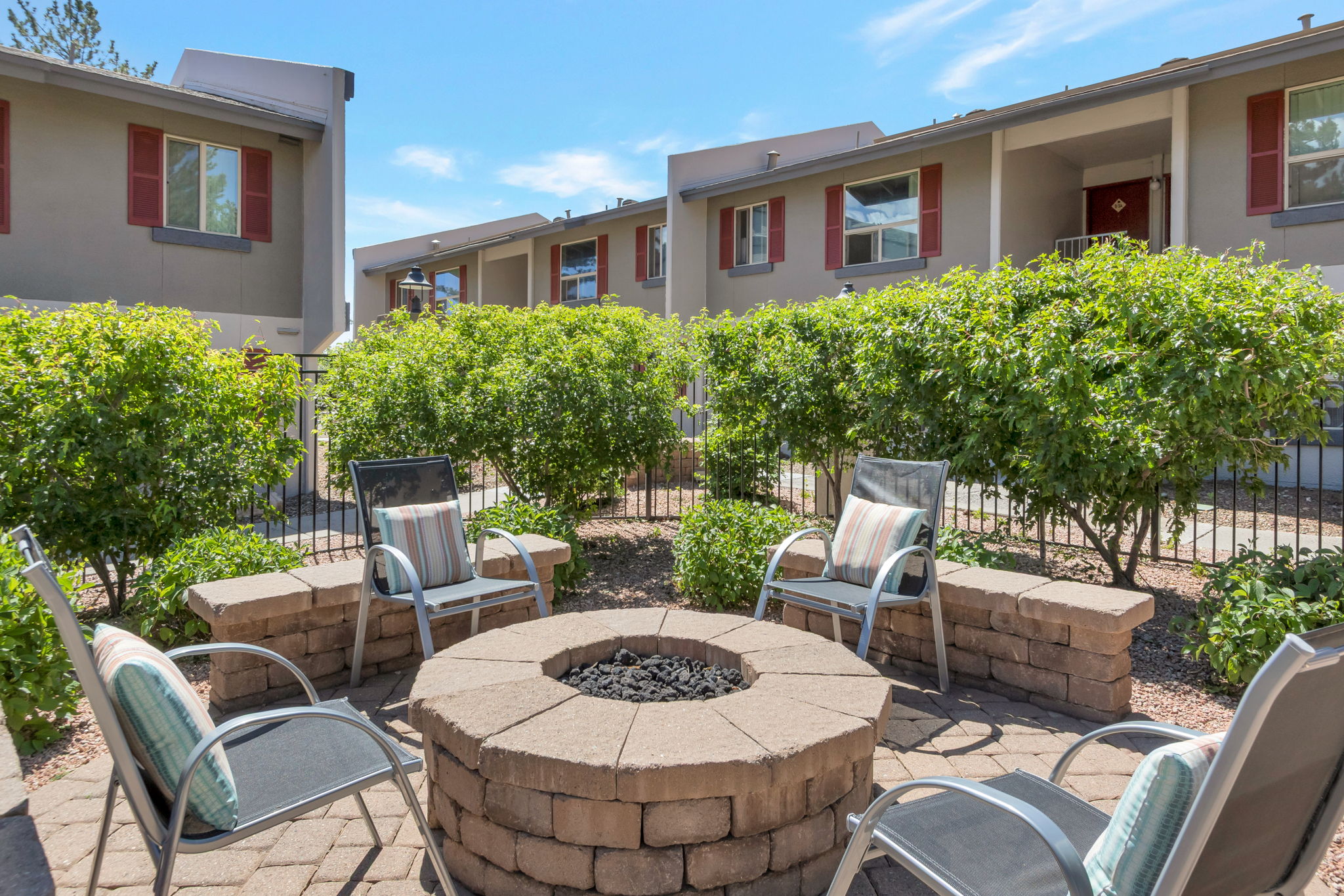 Seating area with four metal chairs around a circular stone fire pit, surrounded by green bushes and landscaping. In the background, there are apartment buildings with red window shutters under a clear blue sky.