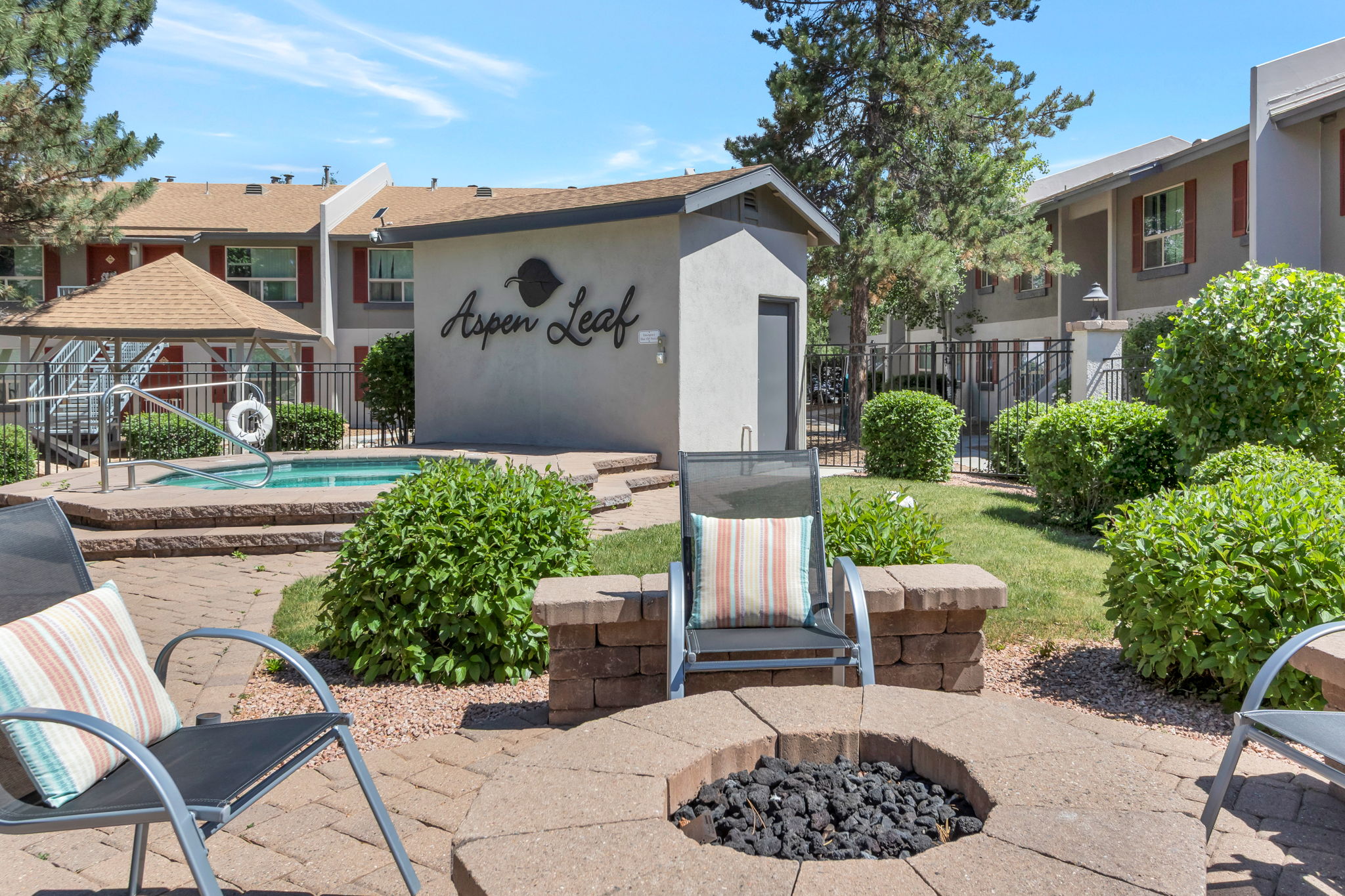 Outdoor patio area featuring a small fire pit surrounded by stone seating, lounge chairs with striped cushions, and a hot tub in the background. The “Aspen Leaf” sign is visible on a nearby building, with manicured bushes and trees adding greenery to the scene. Bright blue sky enhances the inviting atmosphere.