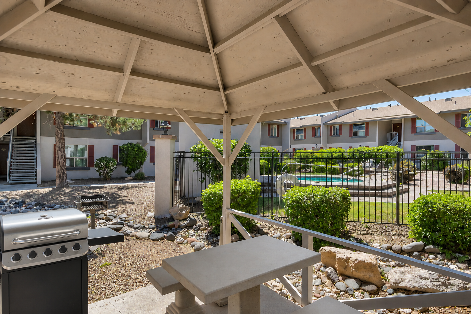 View from a shaded patio with a barbecue grill and picnic table, overlooking a landscaped courtyard featuring a swimming pool and decorative stone accents. Surrounding greenery and an apartment building are visible in the background, creating a relaxed outdoor atmosphere.