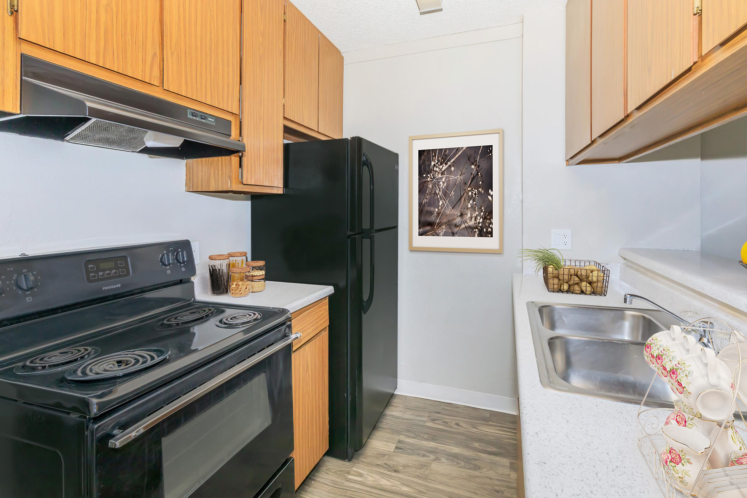 A compact kitchen featuring wooden cabinetry, a black refrigerator, and a black stove with four burners. The countertops are light-colored with a double sink. A framed image of soft lights decorates the wall, and there are decorative elements on the counter, including jars and dishes.