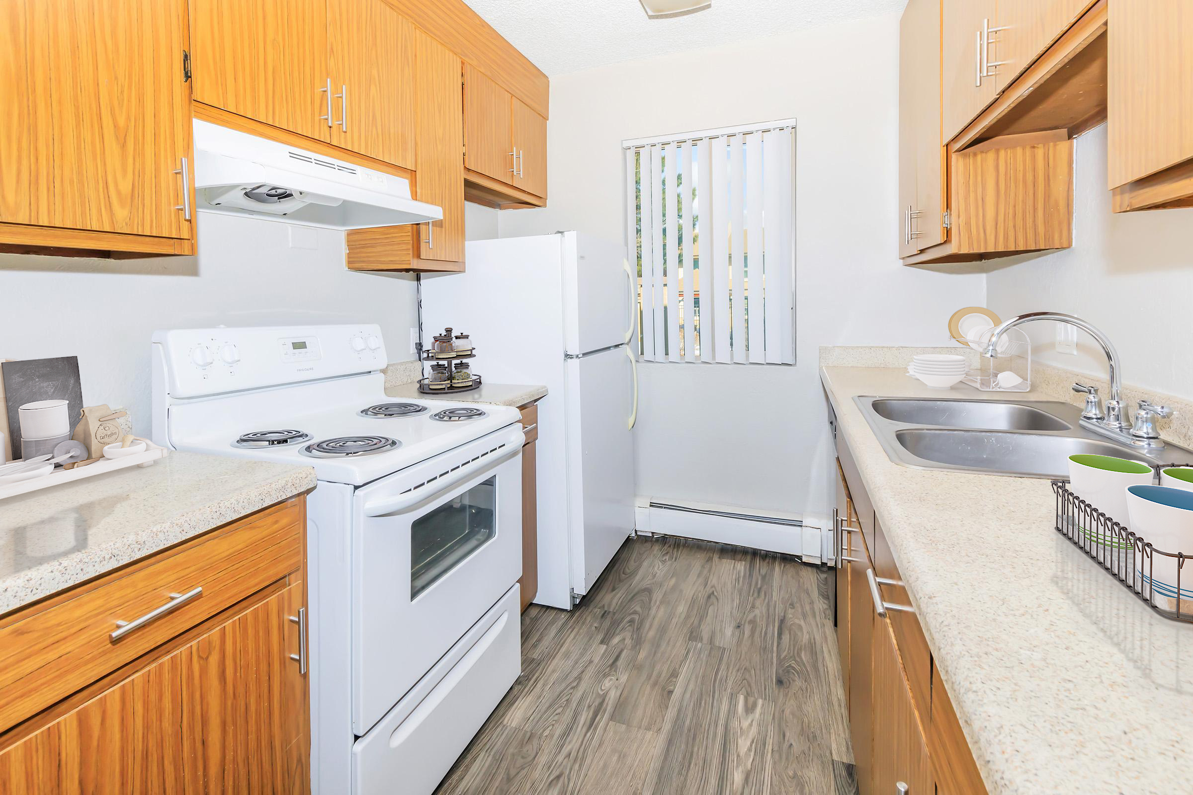 A modern kitchen featuring wooden cabinetry, a white stove and refrigerator, and a double sink. The countertops are light-colored, and the flooring has a wood-like appearance. A window with vertical blinds provides natural light. Kitchen accessories, including dishes, are neatly arranged.