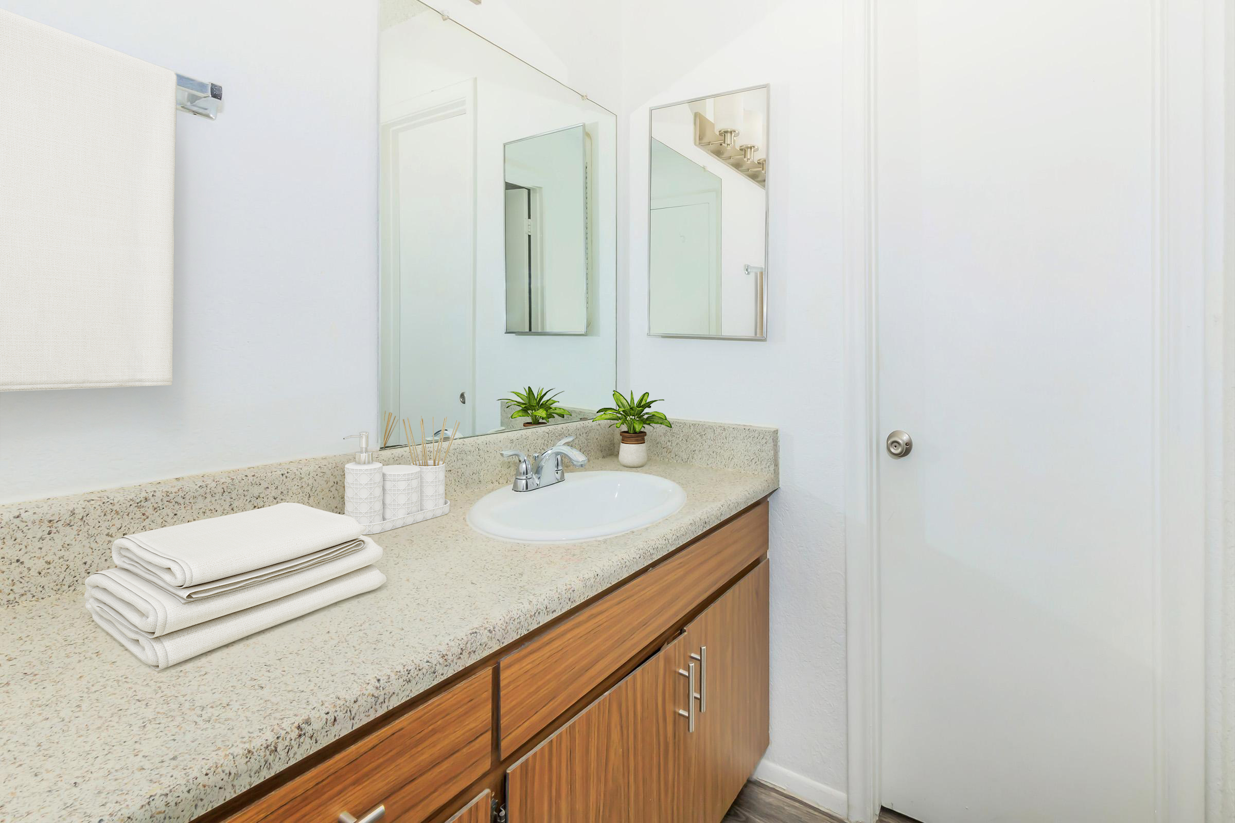 A clean bathroom with a countertop featuring neatly stacked white towels, a small potted plant, and personal care items in containers. The wall is painted white, and there are three mirrors above the sink. The door is partially visible on the right side.