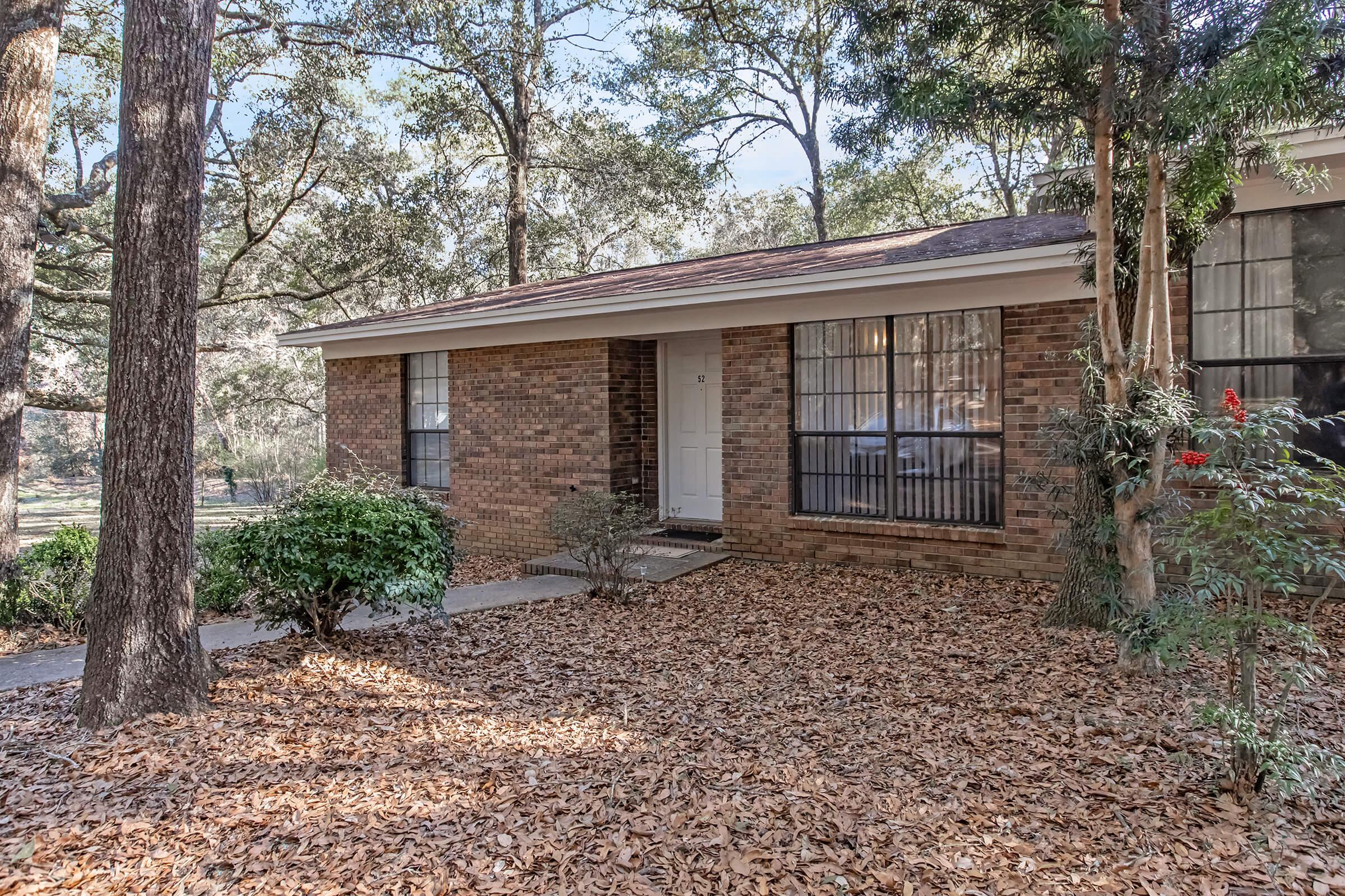 A single-story brick house with large windows, surrounded by trees and scattered fallen leaves. The front yard features a small shrub and a pathway leading to the entrance. Natural light filters through the trees, creating a peaceful atmosphere.