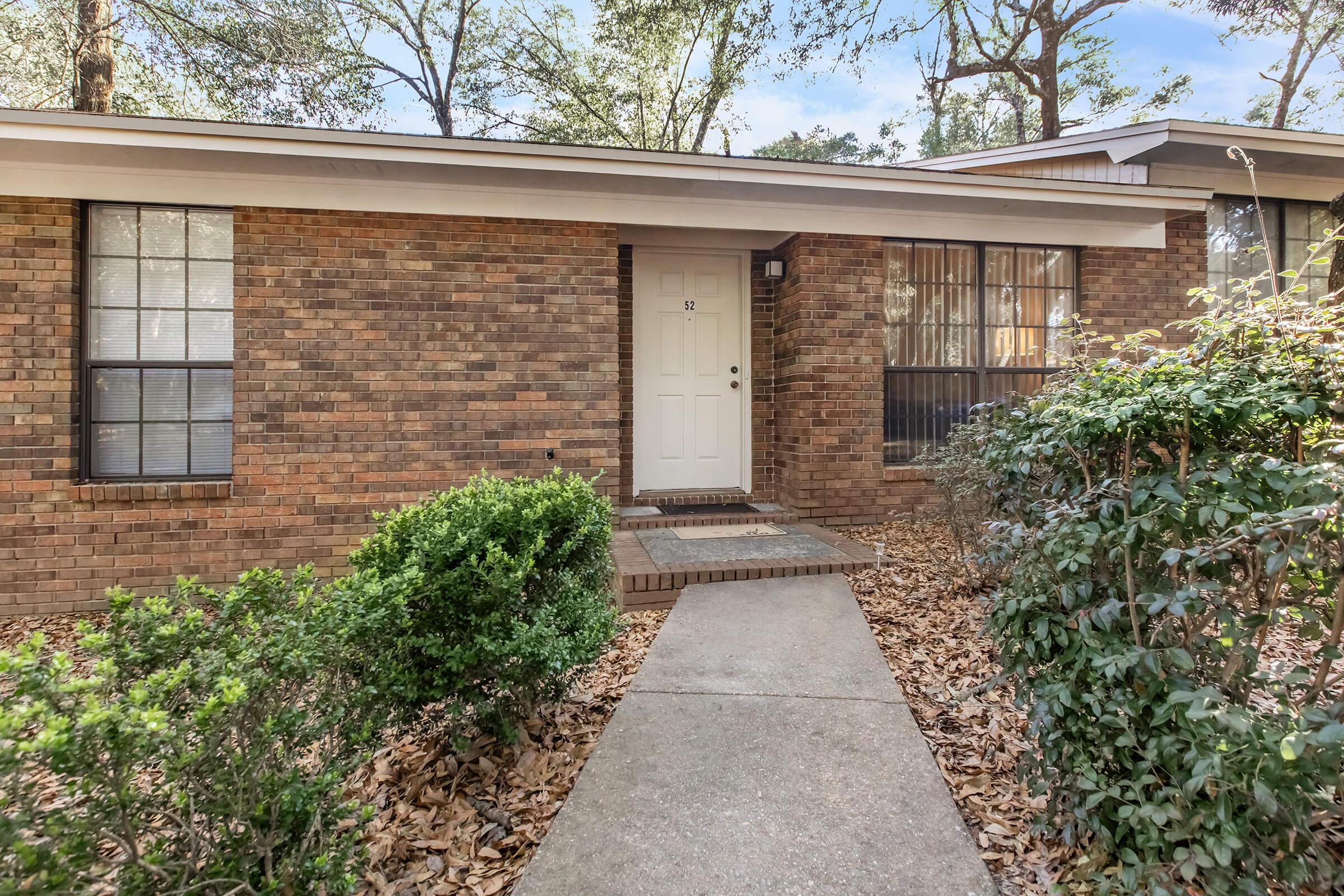 Front view of a brick house with a white door and windows. The walkway is lined with bushes and fallen leaves, leading up to the entrance. Tall trees are visible in the background, indicating a wooded area. The house has a simple, inviting appearance.