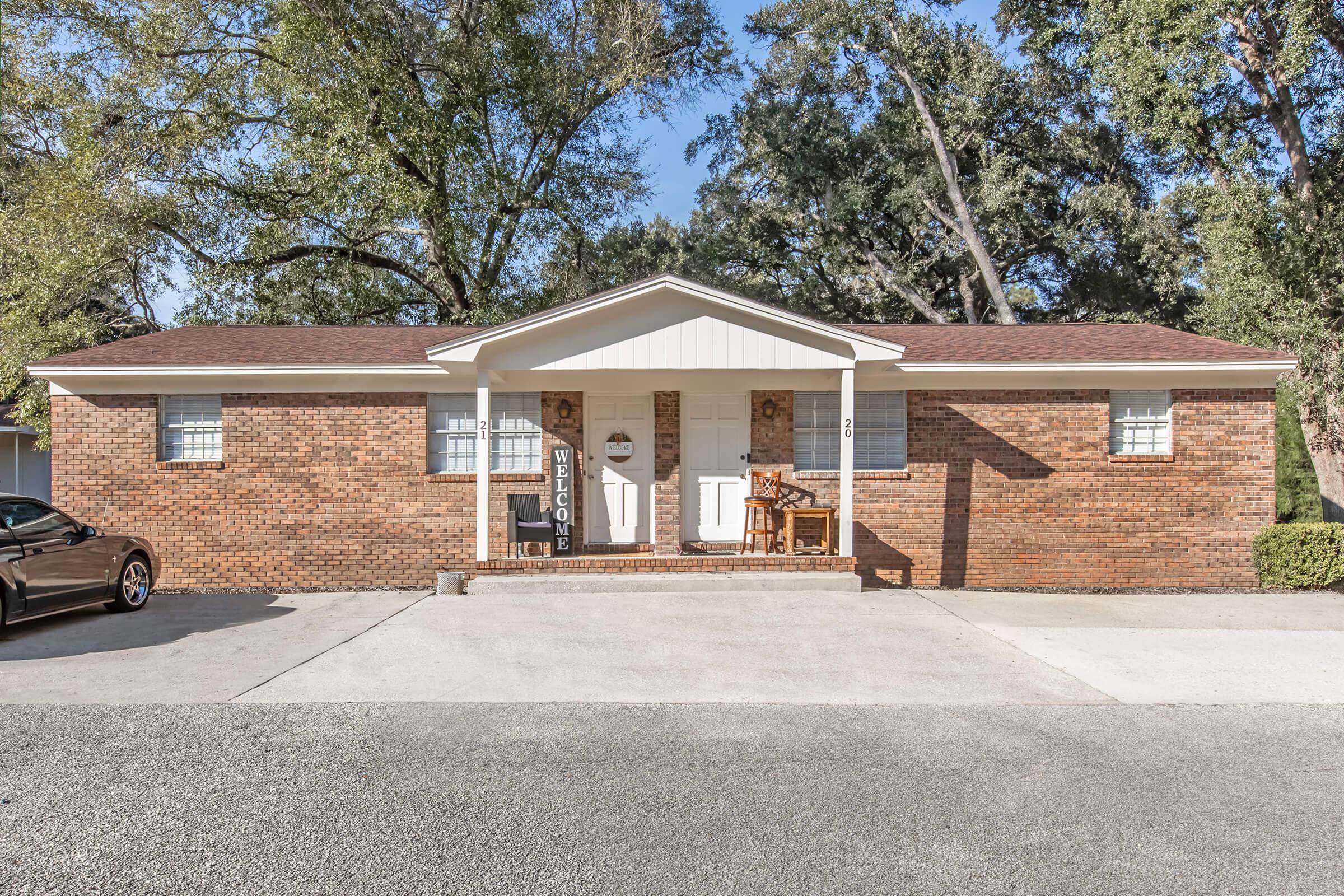 A brick home with a white facade and a gabled roof, featuring a welcoming entrance with a "Welcome" sign. There are two front doors and a small porch with a chair. The driveway is paved, and there are trees and shrubs surrounding the house, creating a pleasant outdoor space.