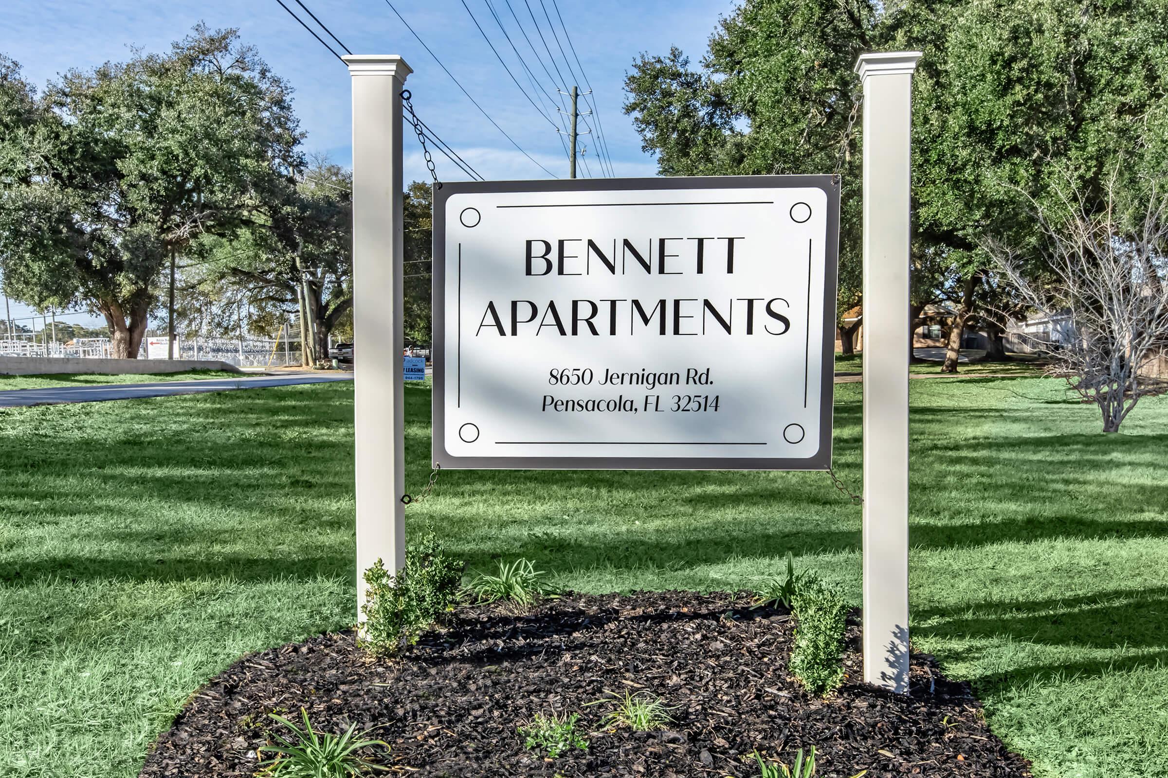 Sign for Bennett Apartments, located at 8650 Jernigan Rd, Pensacola, FL 25314. The sign is white with black lettering and is surrounded by green grass and small plants, with trees in the background.