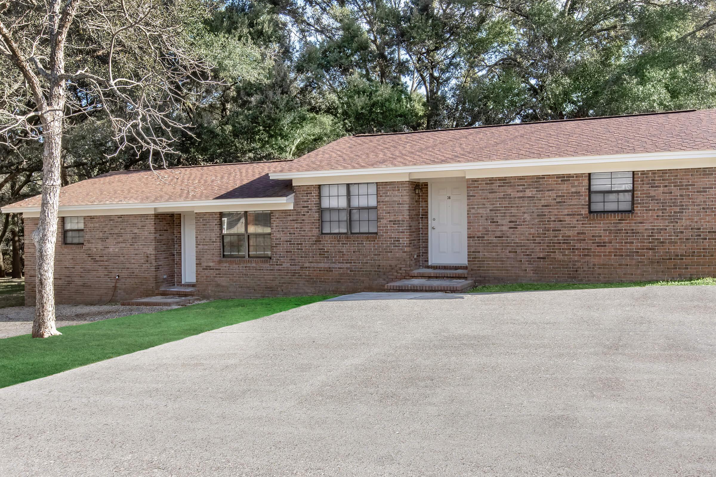 A single-story brick house with a prominent front entrance, surrounded by trees. The driveway is gravel, leading up to the house, which features multiple windows. There's a small yard in front with grass, and the overall setting appears peaceful and suburban.