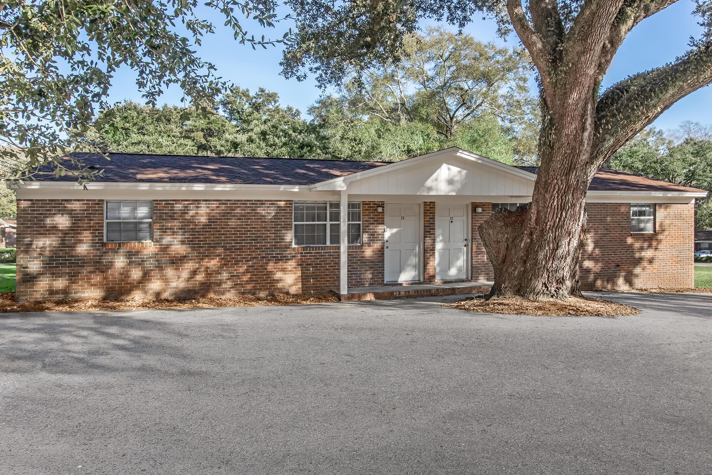 A brick house with a white trim, two front doors, and large windows, set in a grassy area. A large tree is positioned near the entrance, and the driveway is made of gravel. The surrounding landscape features green grass and trees.