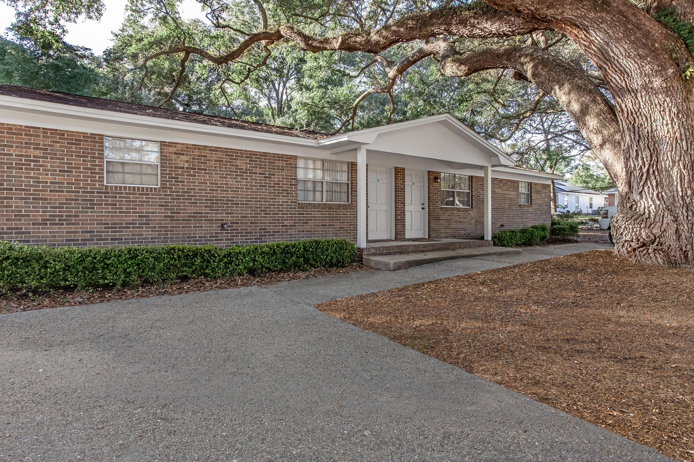 A single-story brick house with white trim and a welcoming front porch, surrounded by lush green shrubs. A large tree with a thick trunk provides shade in the front yard, and a gravel driveway leads up to the entrance.