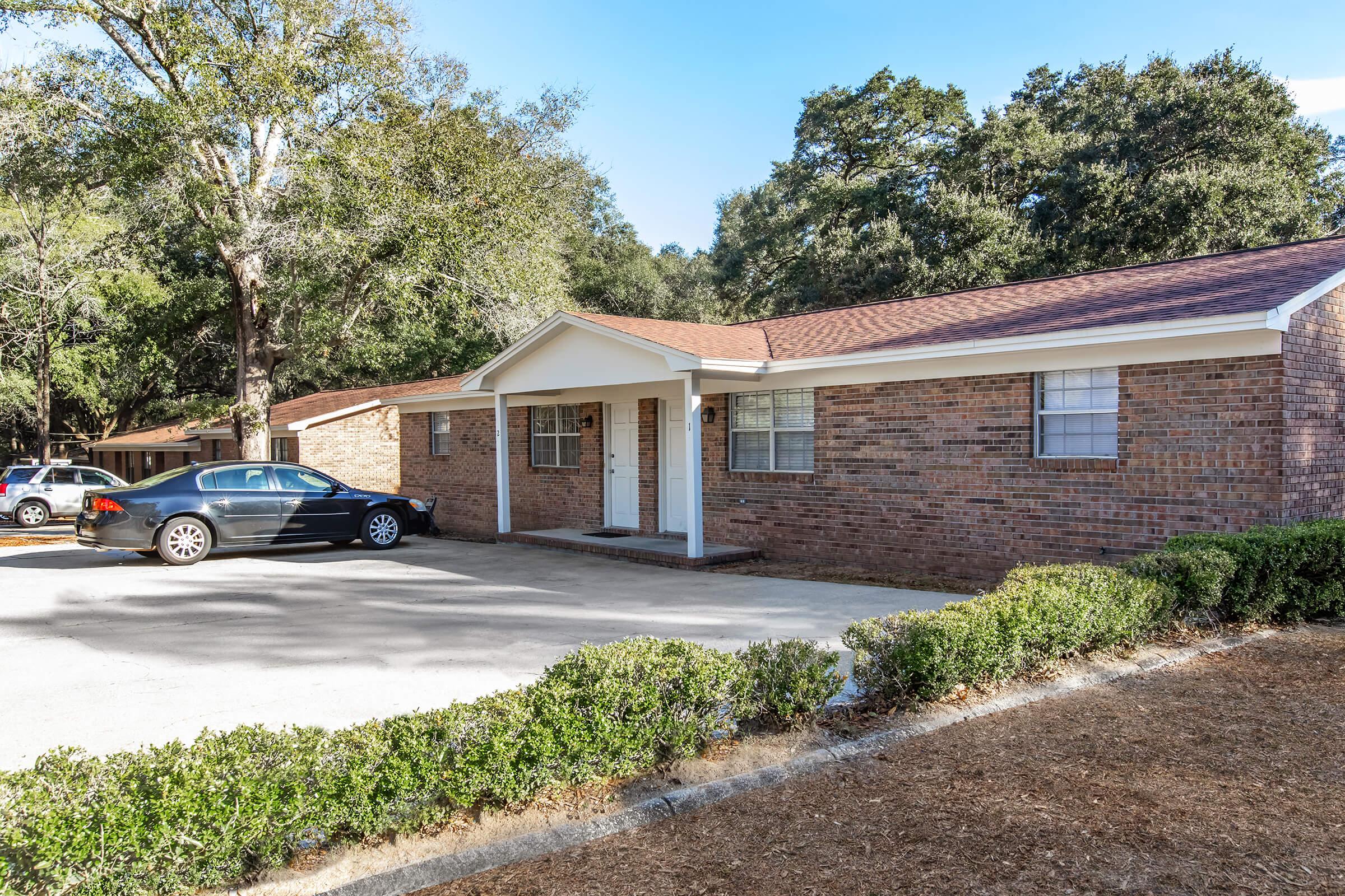 A single-story brick house with a simple front entrance, surrounded by trees and shrubs. A black car is parked in the driveway, and the scene shows a clear sky with no visible people. The house has multiple windows, and a well-maintained yard with gravel.