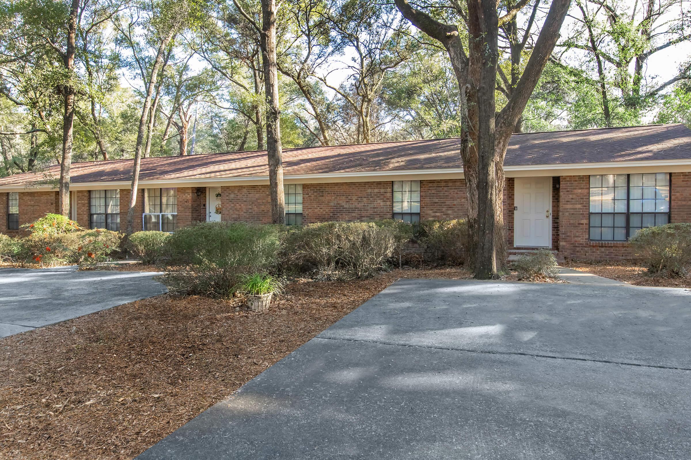 A brick single-story residential building surrounded by trees and shrubs, featuring a driveway with a parking area. The building has multiple windows and a front entrance, all set within a natural, wooded environment. Leaf litter covers the ground, adding to the serene outdoor setting.