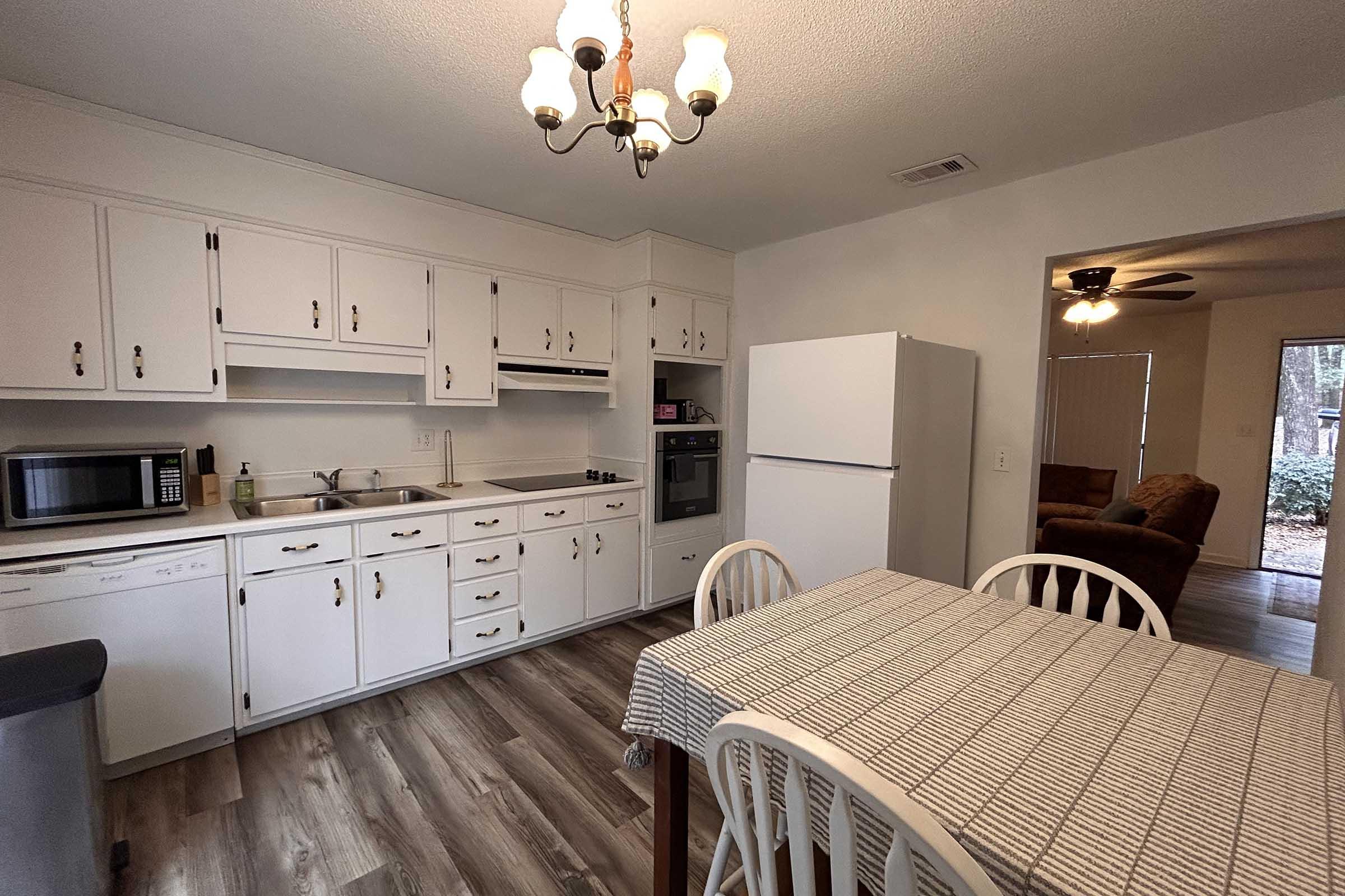 A kitchen with white cabinets, a countertop, and a dining table set for four. The kitchen features a microwave, a stove, and a refrigerator. A doorway leads to a living area with a couch. The flooring is wooden, and there's a chandelier hanging from the ceiling.