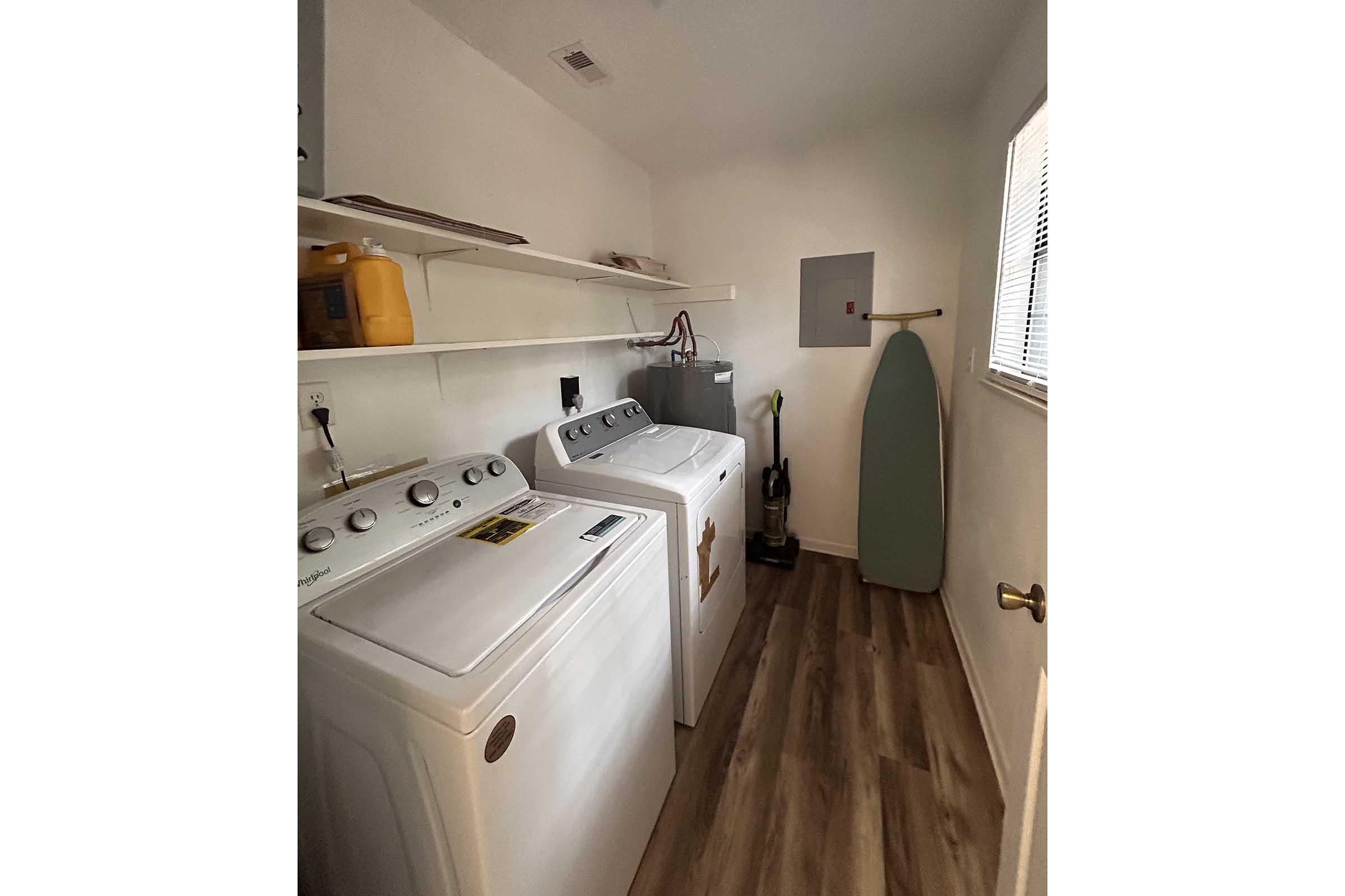 A laundry room featuring two white washing machines side by side, shelves above with cleaning supplies, a green ironing board leaning against the wall, and a small window allowing natural light. The floor is made of light-colored wood.