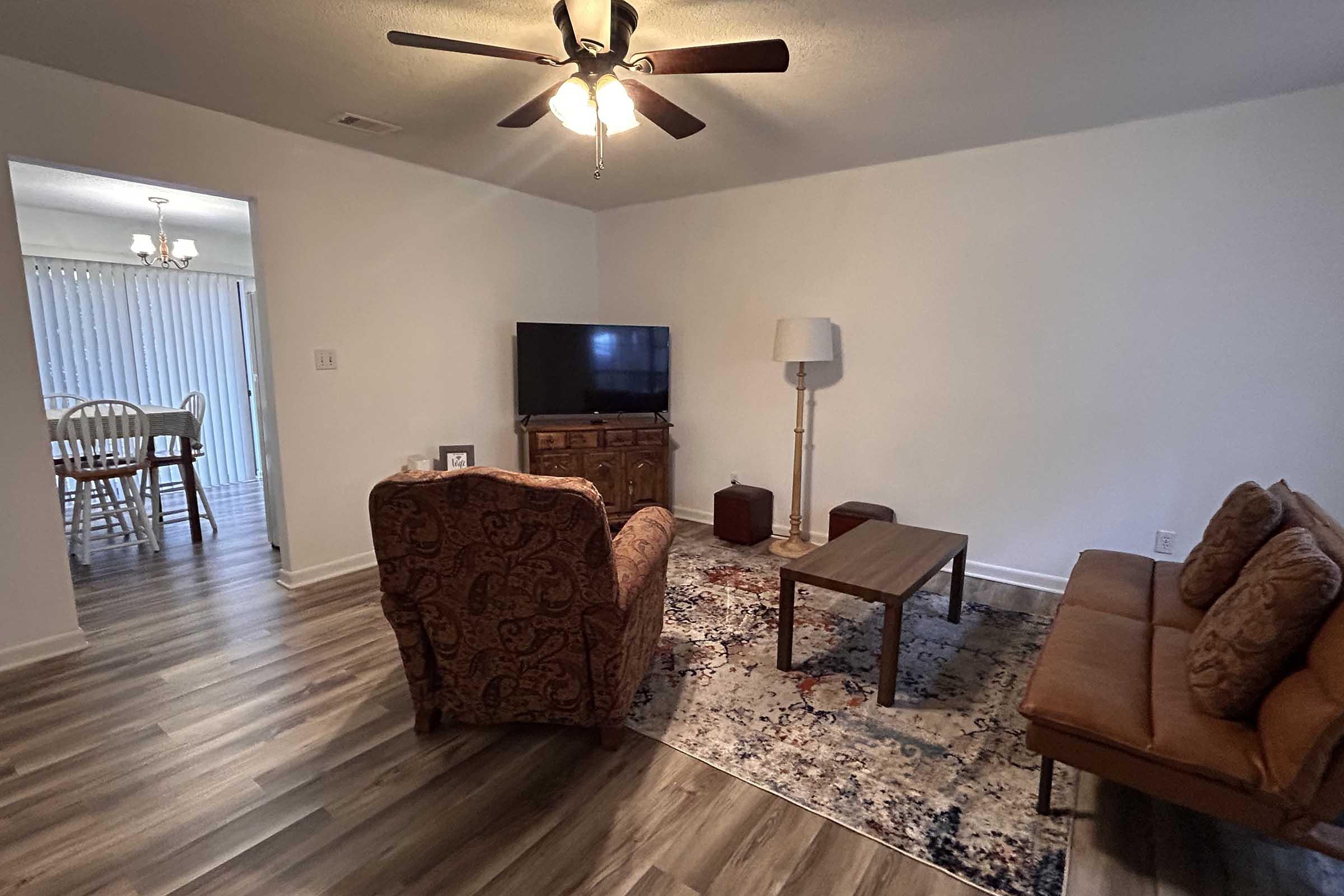 A cozy living room with a patterned rug, a wooden coffee table, a brown sofa, an armchair, a TV on a stand, and a lamp. There is a doorway leading to another room with a dining area visible in the background.