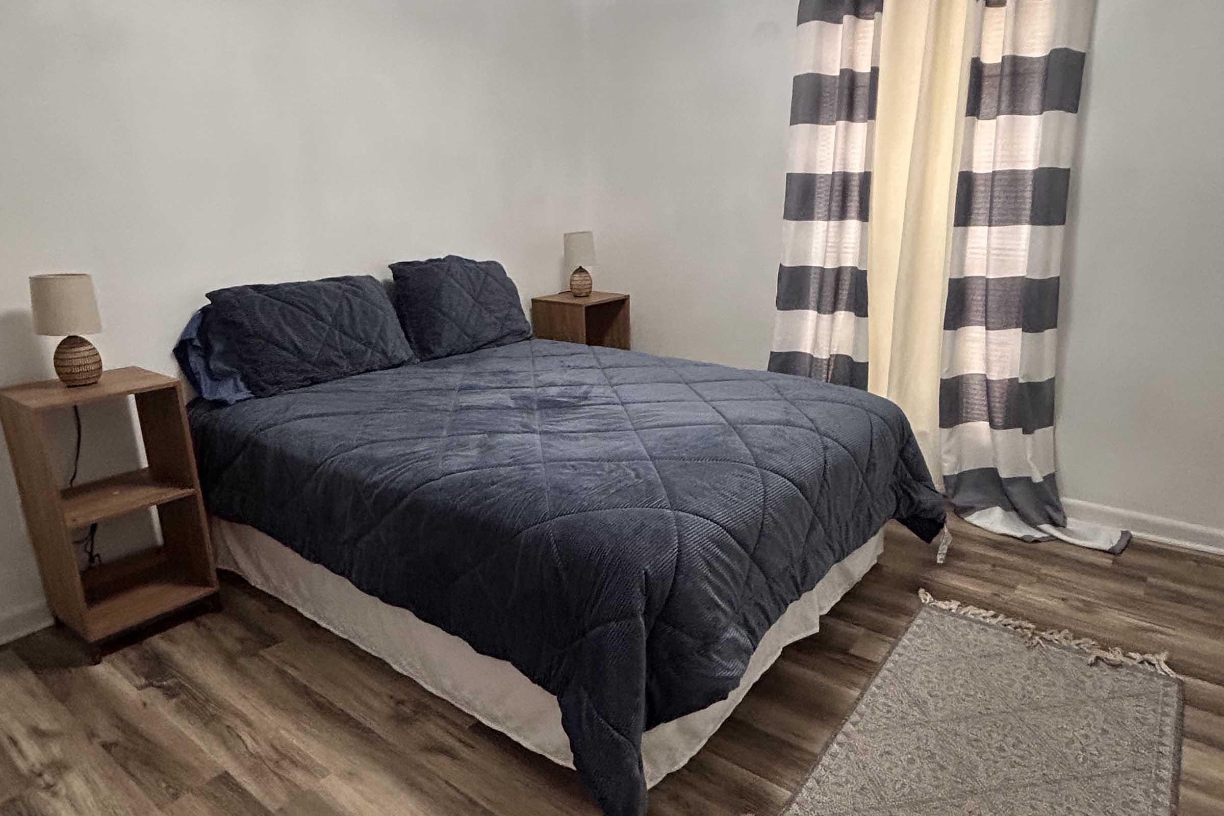 A cozy bedroom featuring a neatly made bed with a navy blue quilt and two matching pillows. Flanking the bed are two wooden nightstands with lamps. The room has striped curtains that let in natural light, and a light-colored rug is visible on the floor.