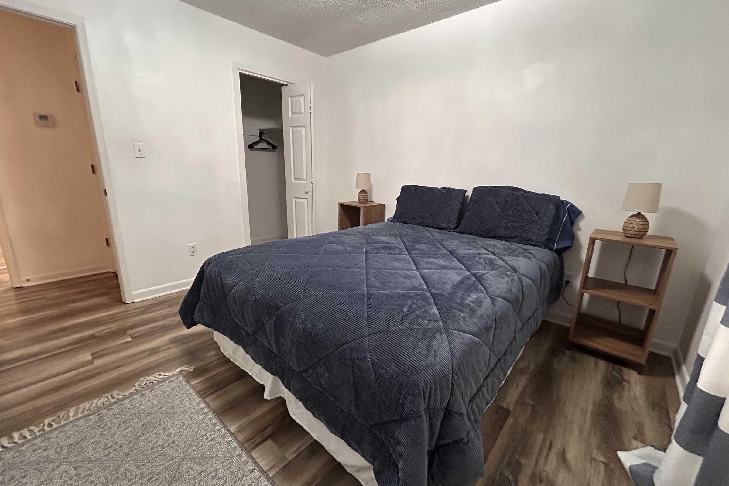 A cozy bedroom featuring a neatly made bed with blue bedding, a nightstand with a lamp on one side, and a closet visible in the background. The walls are painted white, and the floor has wooden planks, creating a warm and inviting atmosphere.