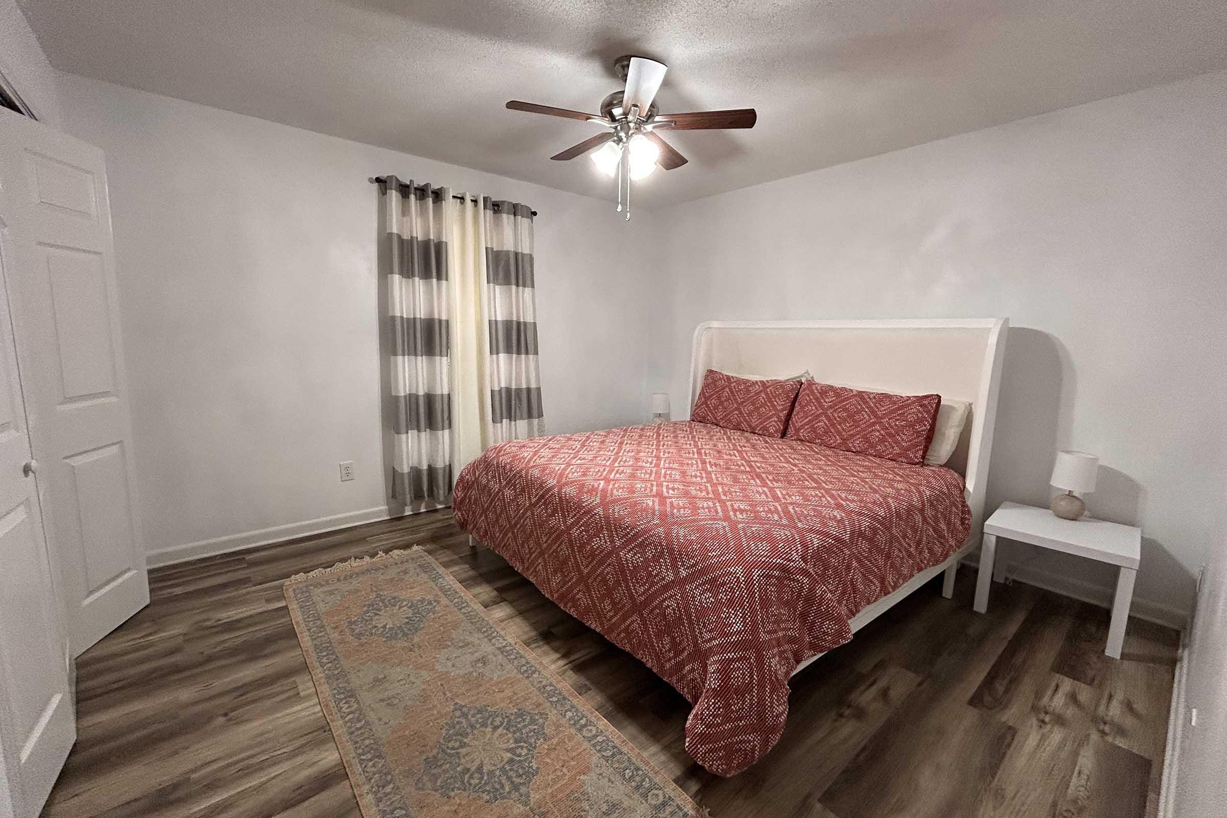 A well-decorated bedroom featuring a white bed with a red patterned comforter, two matching pillows, wooden flooring, a small nightstand with a lamp, a ceiling fan, and large striped curtains.