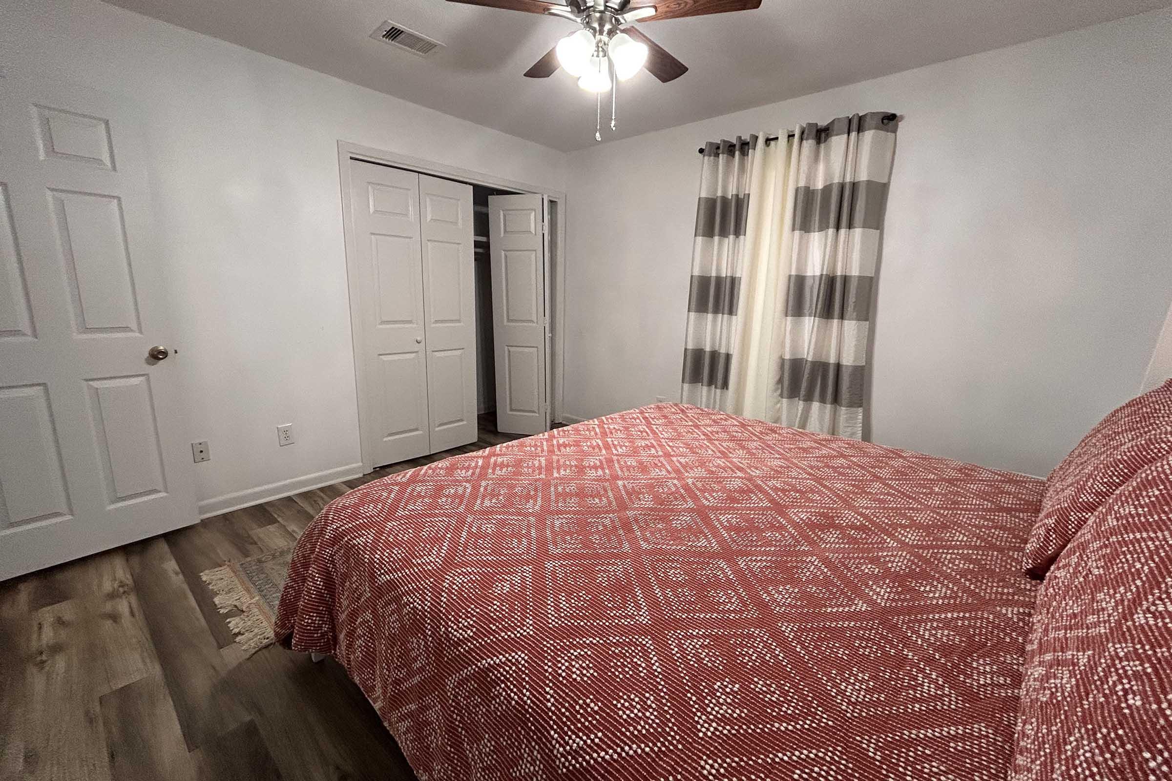 A cozy bedroom featuring a red patterned quilt on a bed, a ceiling fan, and grey and white striped curtains. There are two closed doors, one leading to a closet. The floor is covered with laminate wood planks.