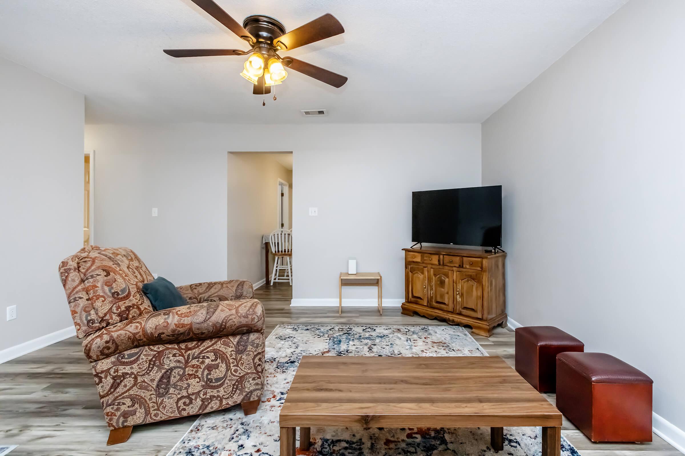 A cozy living room featuring a patterned recliner chair, a wooden coffee table, two small red ottomans, and a TV on a wooden stand. Soft lighting from a ceiling fan creates a warm atmosphere, with neutral walls and flooring that complement the decor. A doorway leads to another room in the background.