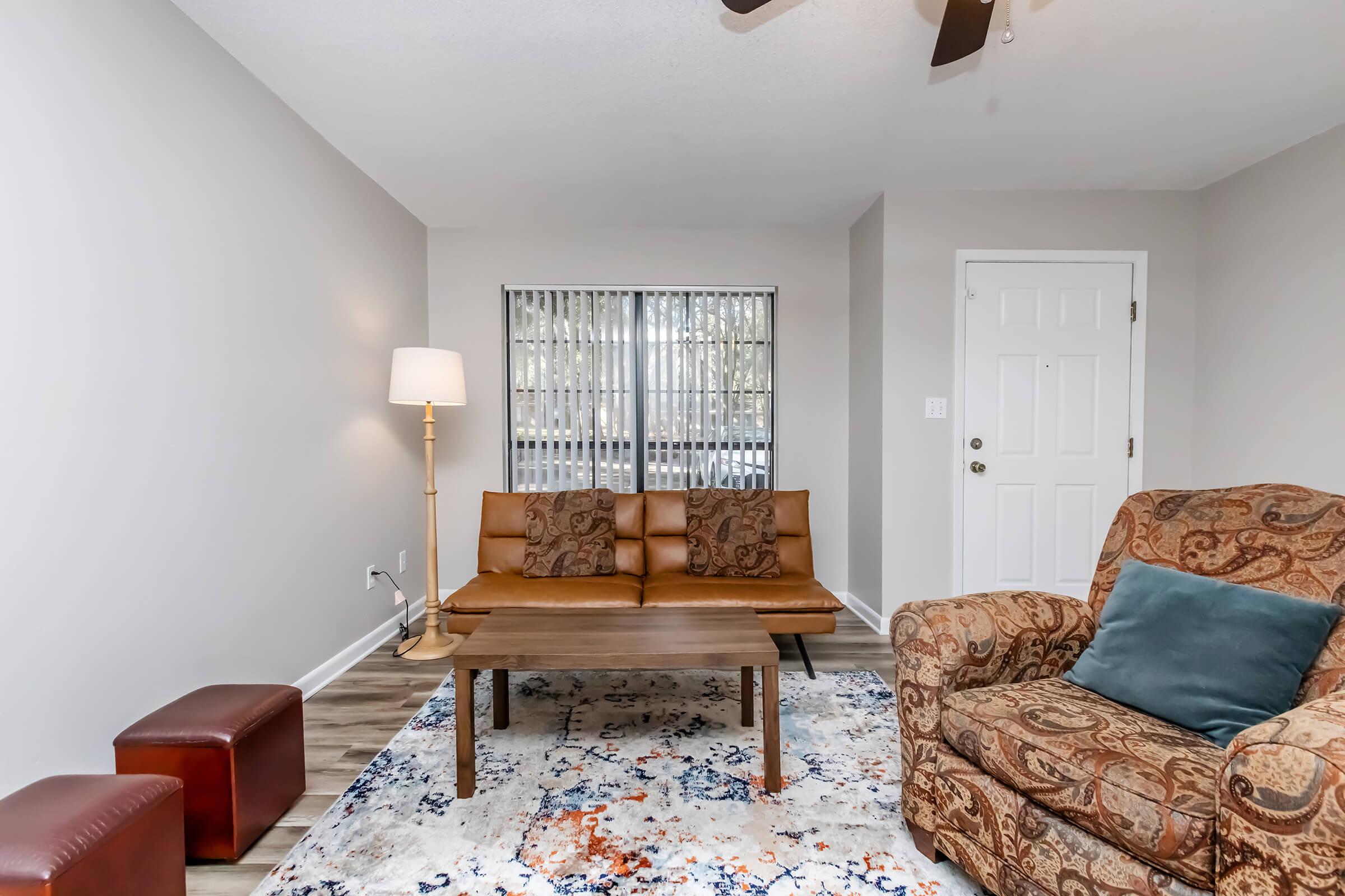 Cozy living room featuring a brown leather sofa and an upholstered armchair. A small table sits in the center on a colorful area rug. A lamp provides soft lighting, and large windows with vertical blinds allow natural light. A front door is visible in the background.