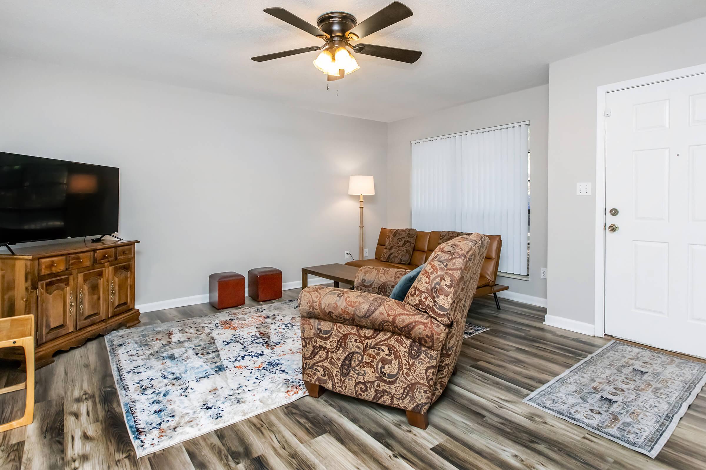 A cozy living room featuring a television, a wooden cabinet, two patterned chairs, a lamp, and a decorative area rug. The walls are painted light gray, and there are wooden floors. A light fixture is visible on the ceiling, and a door leads outside, with vertical blinds on a window.