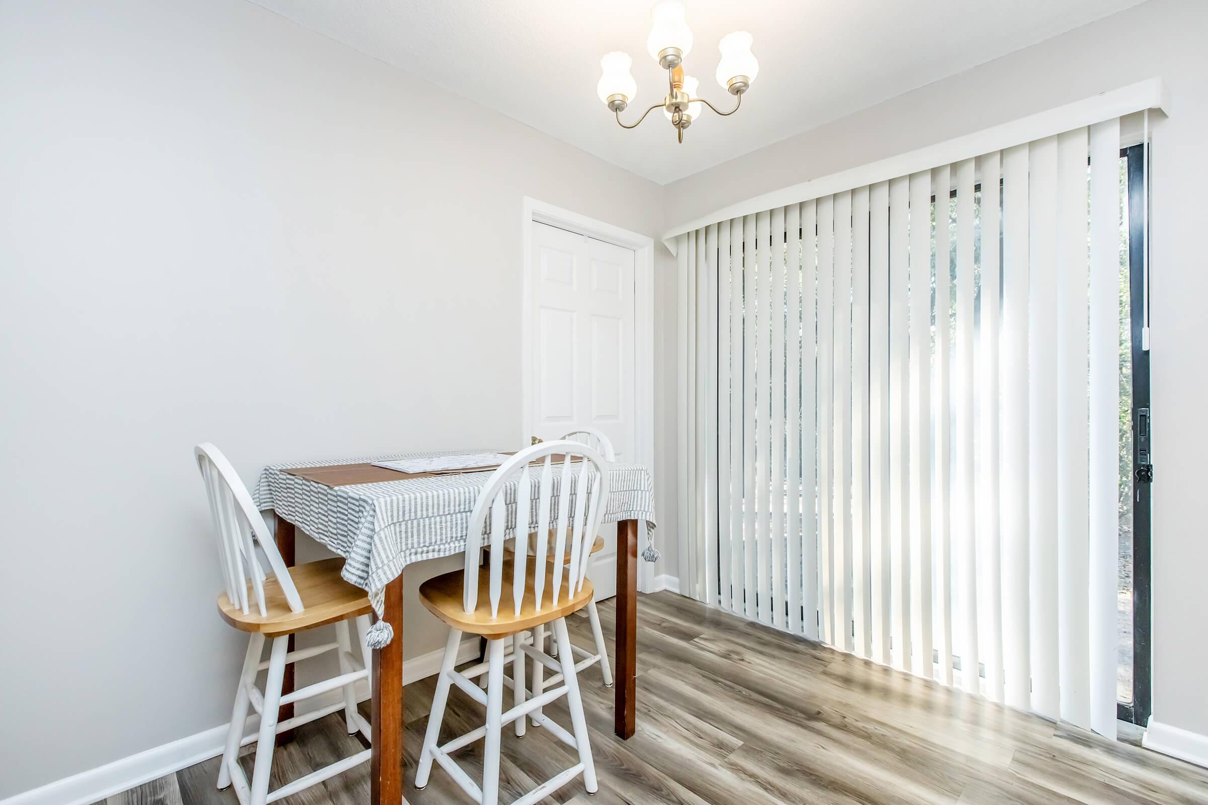 A bright and simple dining area featuring a wooden table covered with a checkered tablecloth and two white chairs. A chandelier hangs from the ceiling, and there are sliding glass doors with vertical blinds letting in natural light. The floor is a light-colored laminate.