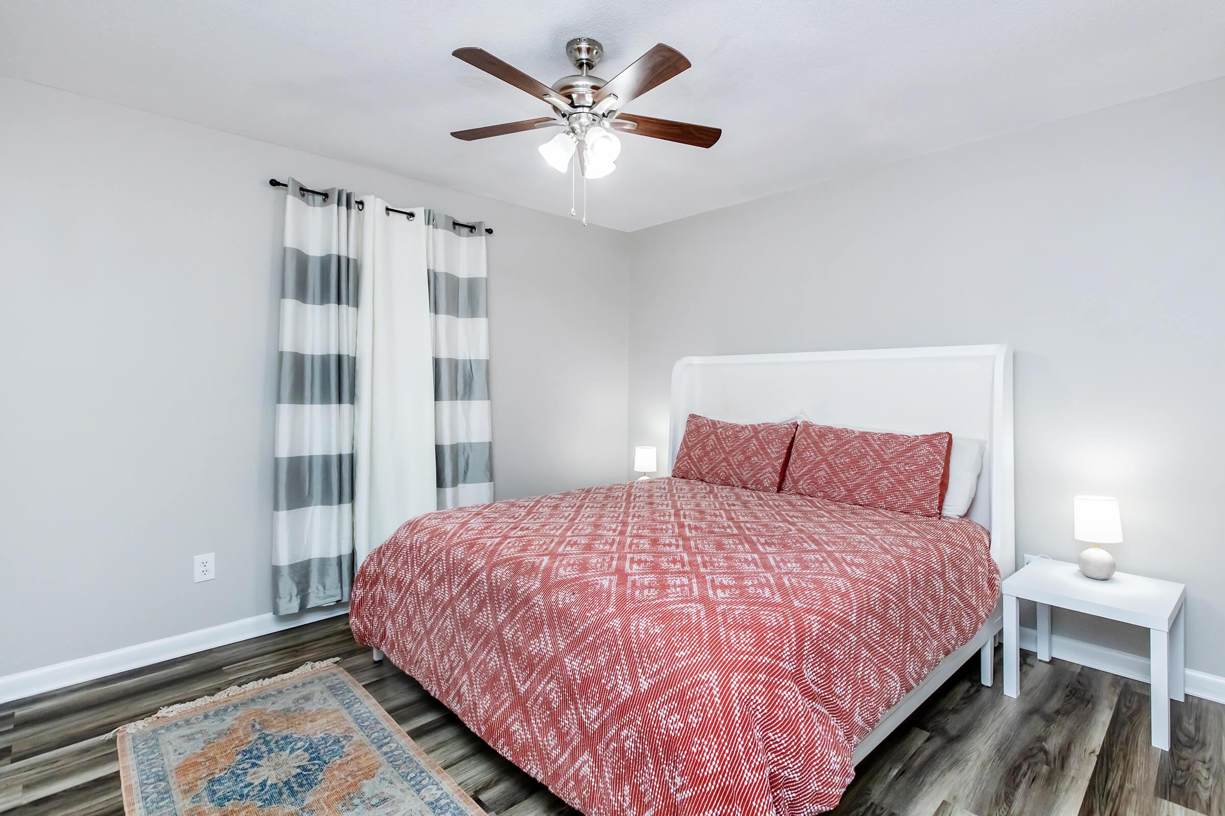 Cozy bedroom featuring a white bed with a red patterned comforter, bedside tables with lamps, a ceiling fan, and gray and white striped curtains. The floor is wooden, and a small area rug adds warmth to the space. The walls are painted a light gray, creating a calm atmosphere.