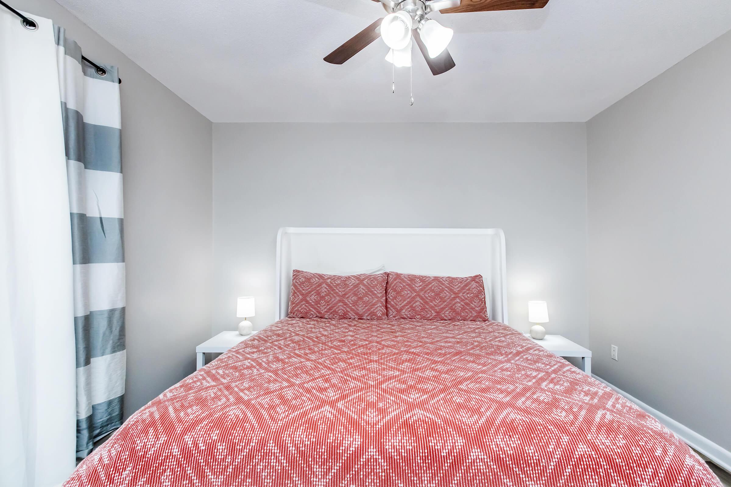 A cozy bedroom featuring a white bed frame with a red patterned comforter. There are two white nightstands with lamp on each side, and gray striped curtains hanging by a window. The walls are painted a light gray, and a ceiling fan with light is installed above.