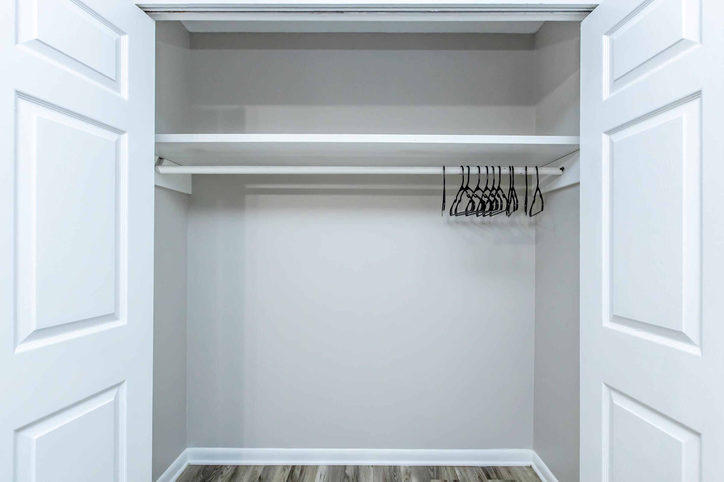 An empty closet with white double doors, featuring a single shelf above and a few empty hangers hanging from the rod. The walls are painted in a light color, and the floor has a wood-like appearance.