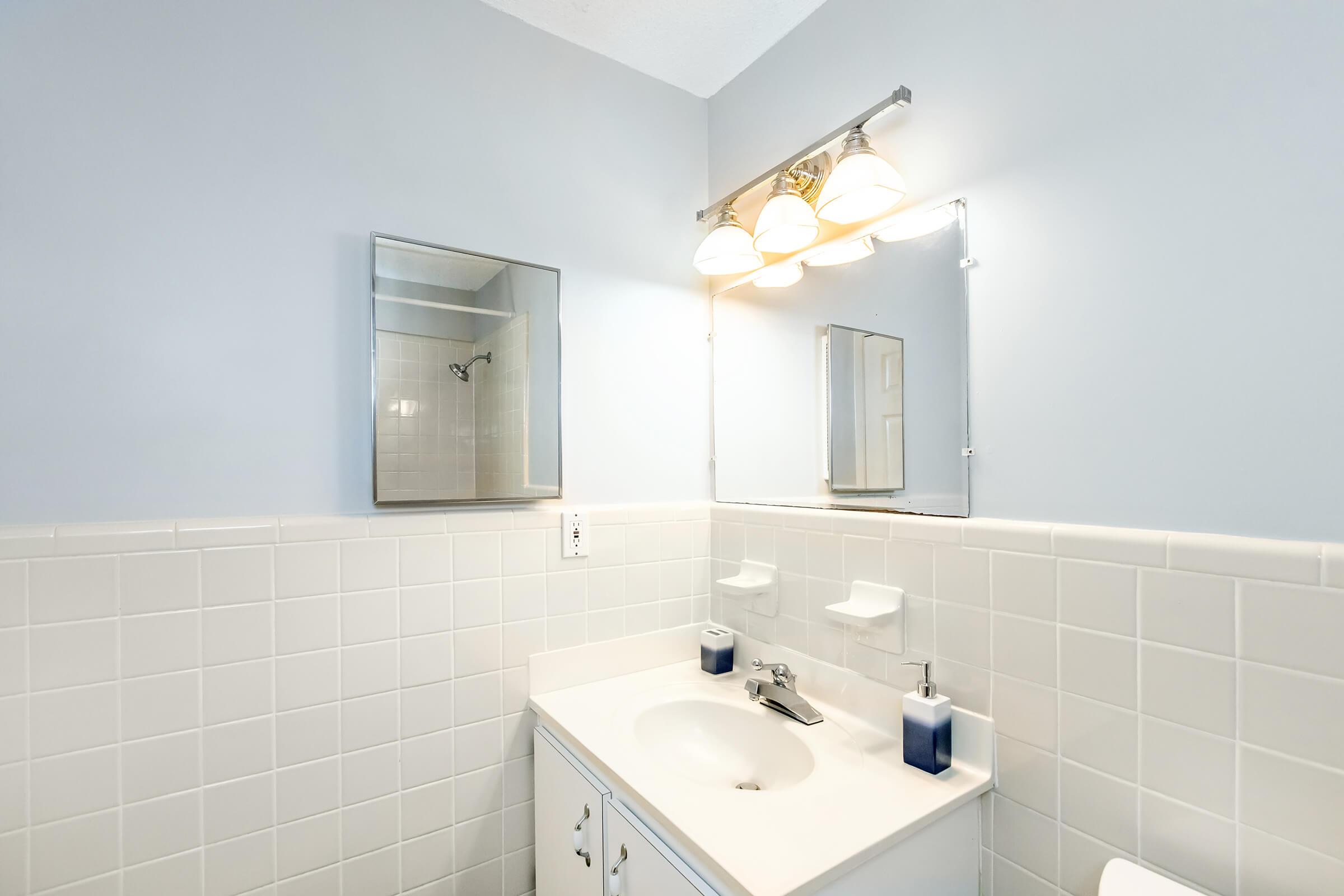 A well-lit bathroom featuring light blue walls, a white tiled backsplash, a sink with a silver faucet, a mirror above the sink, and dual mirrors on either side. The area includes soap dispensers and a shower area visible in the background. The design is clean and modern.