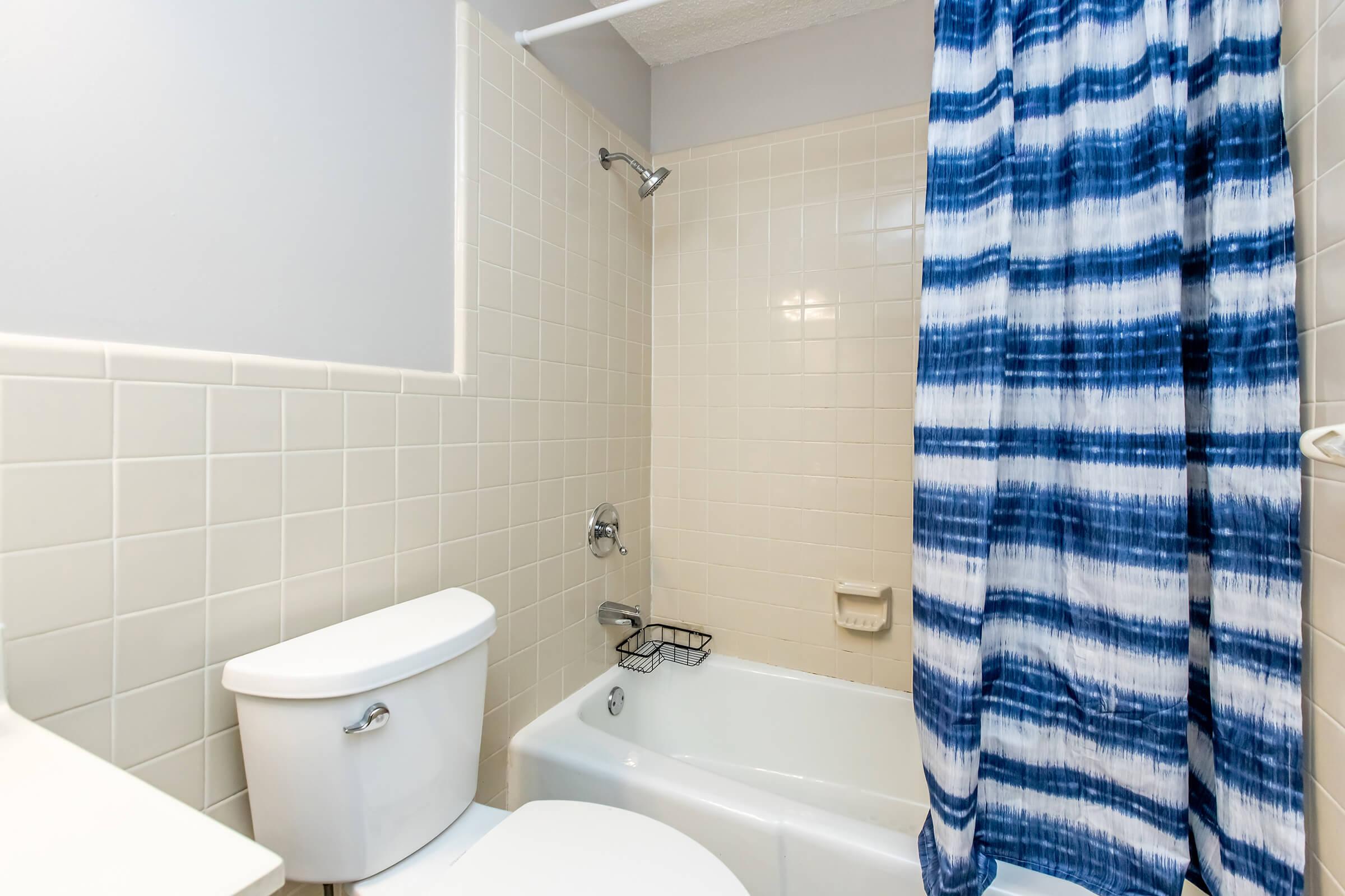 A clean bathroom featuring a white toilet, a bathtub with a blue and white striped shower curtain, tiled walls in neutral colors, and a small storage basket attached to the wall near the tub. The overall design is simple and functional.