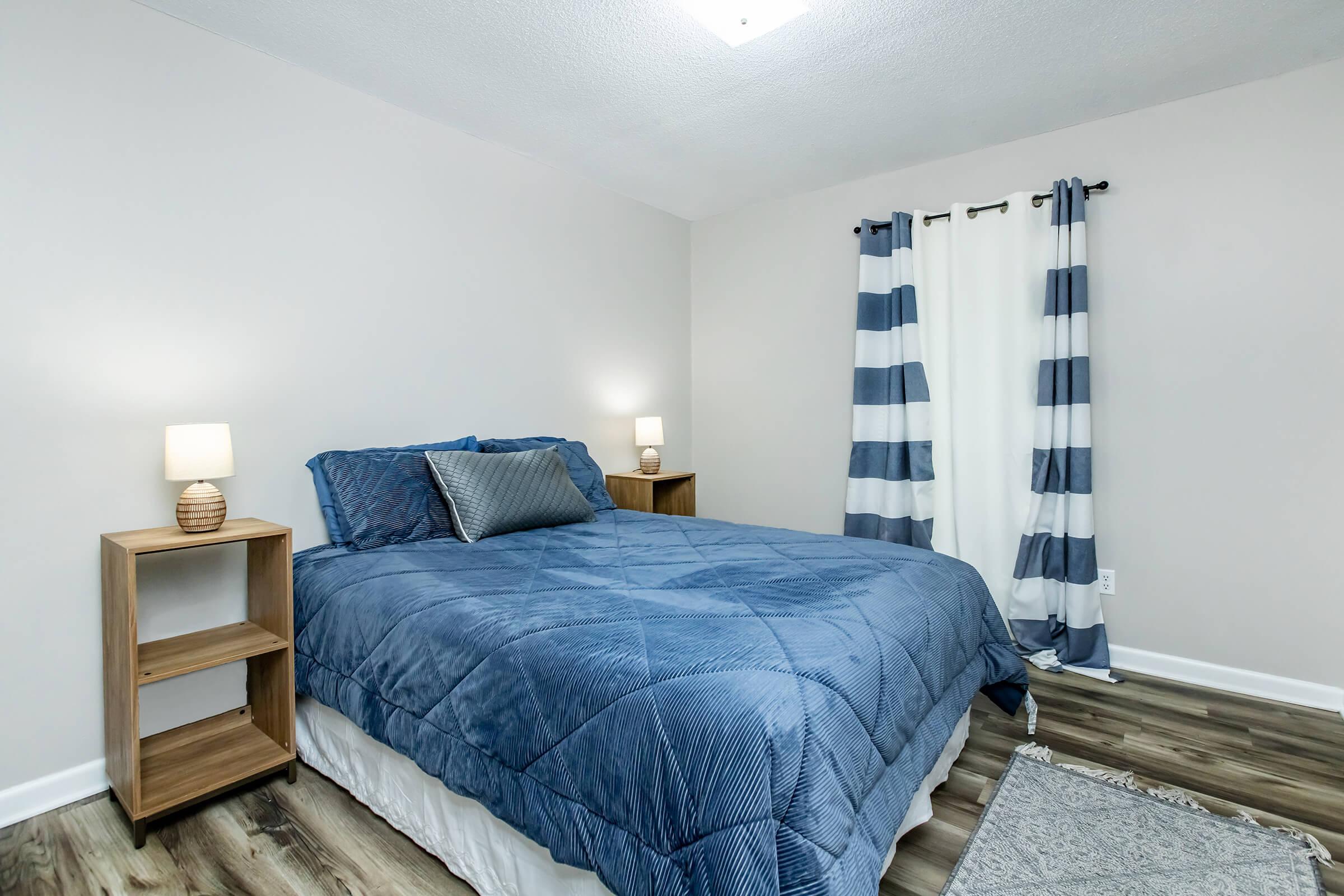 A cozy bedroom featuring a large blue bedspread, two bedside tables with lamps, and striped curtains. The walls are painted a light color, and the floor is covered with a soft rug. The space is well-lit, creating a comfortable and inviting atmosphere.