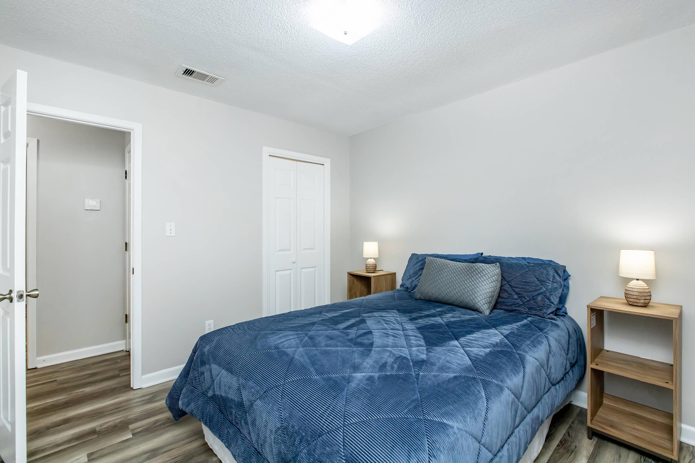 A cozy bedroom featuring a queen-sized bed with a blue quilt, two bedside tables with lamps, and light gray walls. A door leads to another room, and the floor is covered with wood-like laminate. The soft lighting creates a welcoming atmosphere.