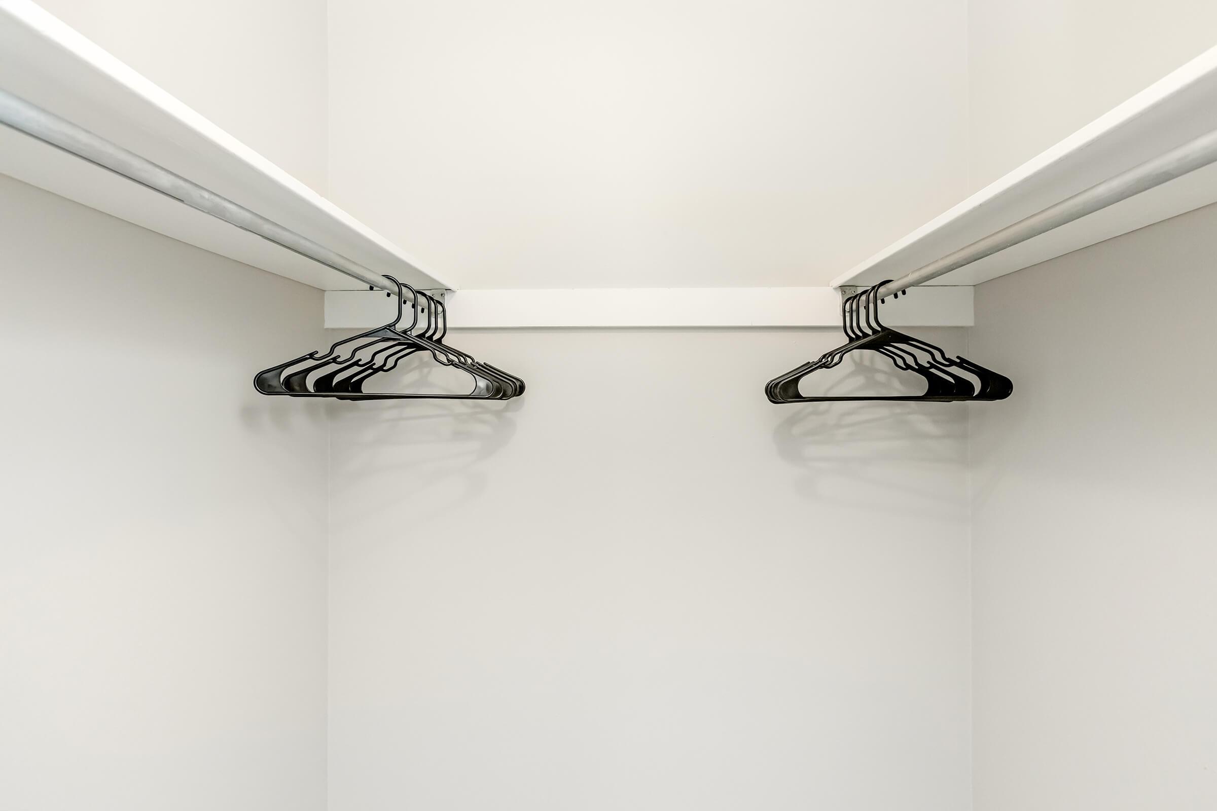 A minimalistic closet featuring two shelves and several black hangers hanging from a white bar. The walls are plain and light-colored, emphasizing the emptiness of the space. Shadows of the hangers are visible on the wall, creating a simplistic and tidy appearance.