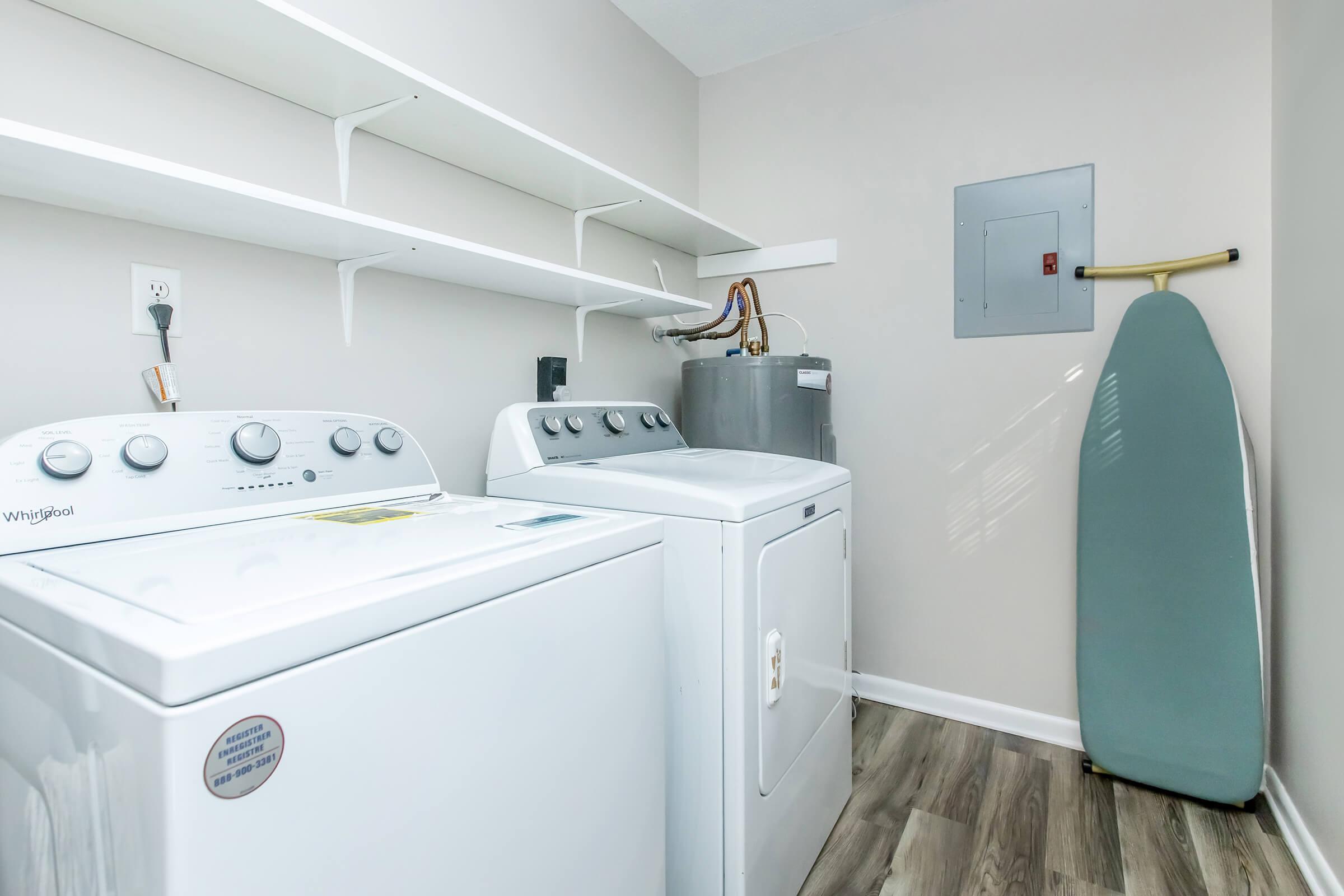 A clean laundry room featuring two white washing machines side by side. Above them are white shelves. To the right, there is a metallic laundry utility sink. A gray ironing board leans against the wall, and a small electrical panel is mounted nearby. The floor is made of wood-like laminate.