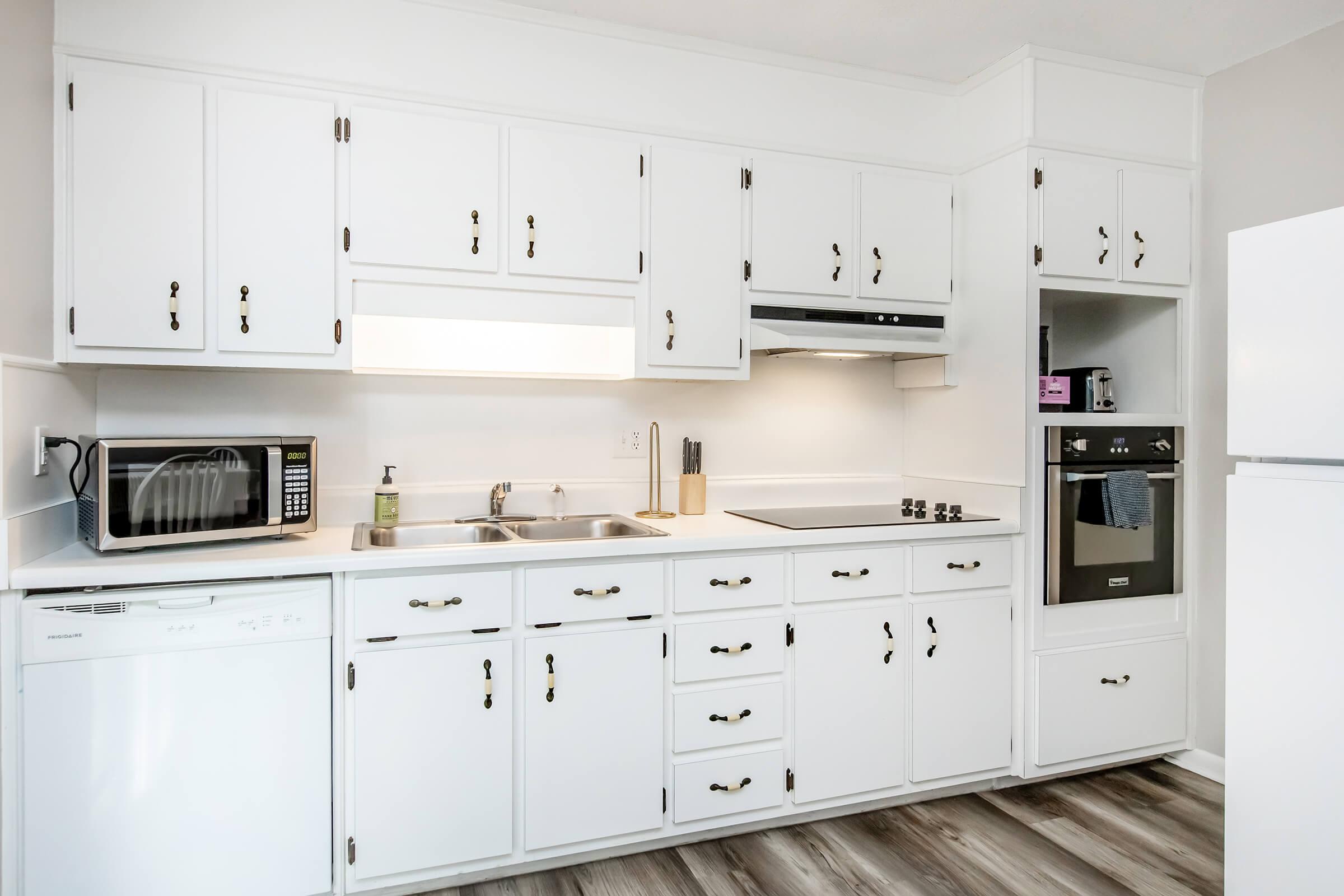 A modern kitchen featuring white cabinets, stainless steel appliances including a microwave, oven, and dishwasher. The kitchen has a double sink and a smooth cooktop, complemented by a neutral-colored wall and wood-style flooring, creating a bright and inviting atmosphere.