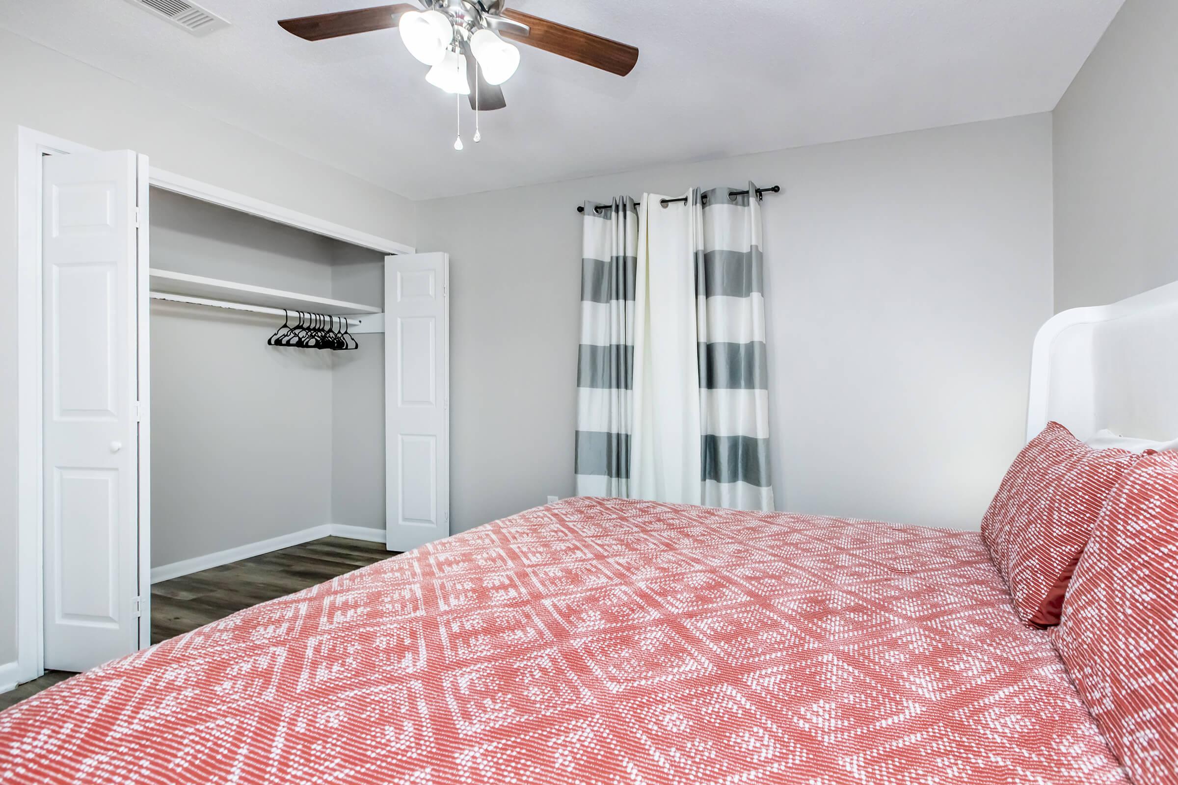 A well-lit bedroom featuring a neatly made red patterned bedspread. In the background, there is a closet with open double doors displaying hangers. The room has gray walls, light-colored curtains with stripes, and a ceiling fan, creating a modern and inviting atmosphere.