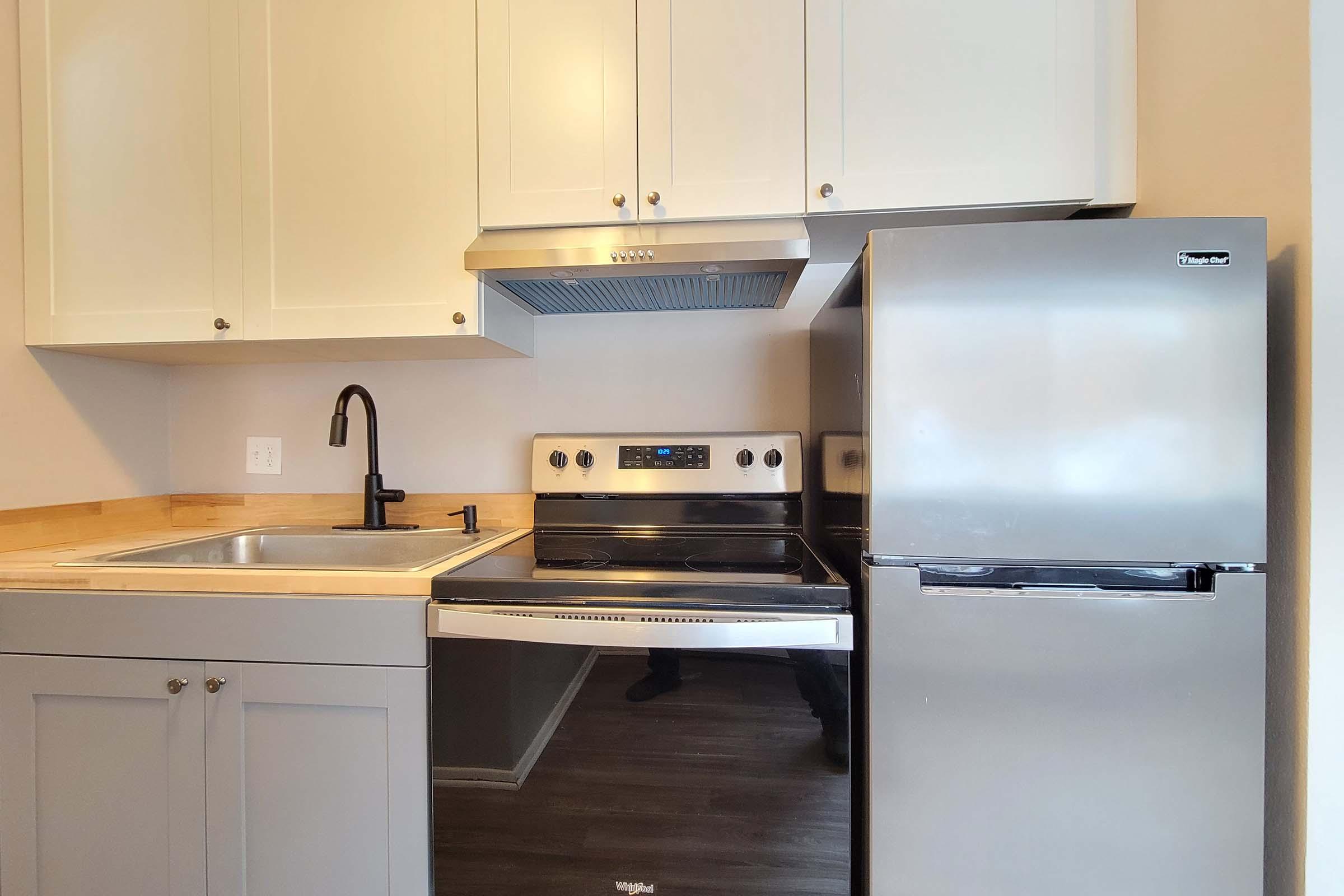 Modern kitchen featuring light gray cabinets, a stainless steel refrigerator, an electric oven and cooktop, and a sleek black faucet above a stainless steel sink. The countertop is wooden, and the space has neutral-colored walls, creating a bright and clean atmosphere.