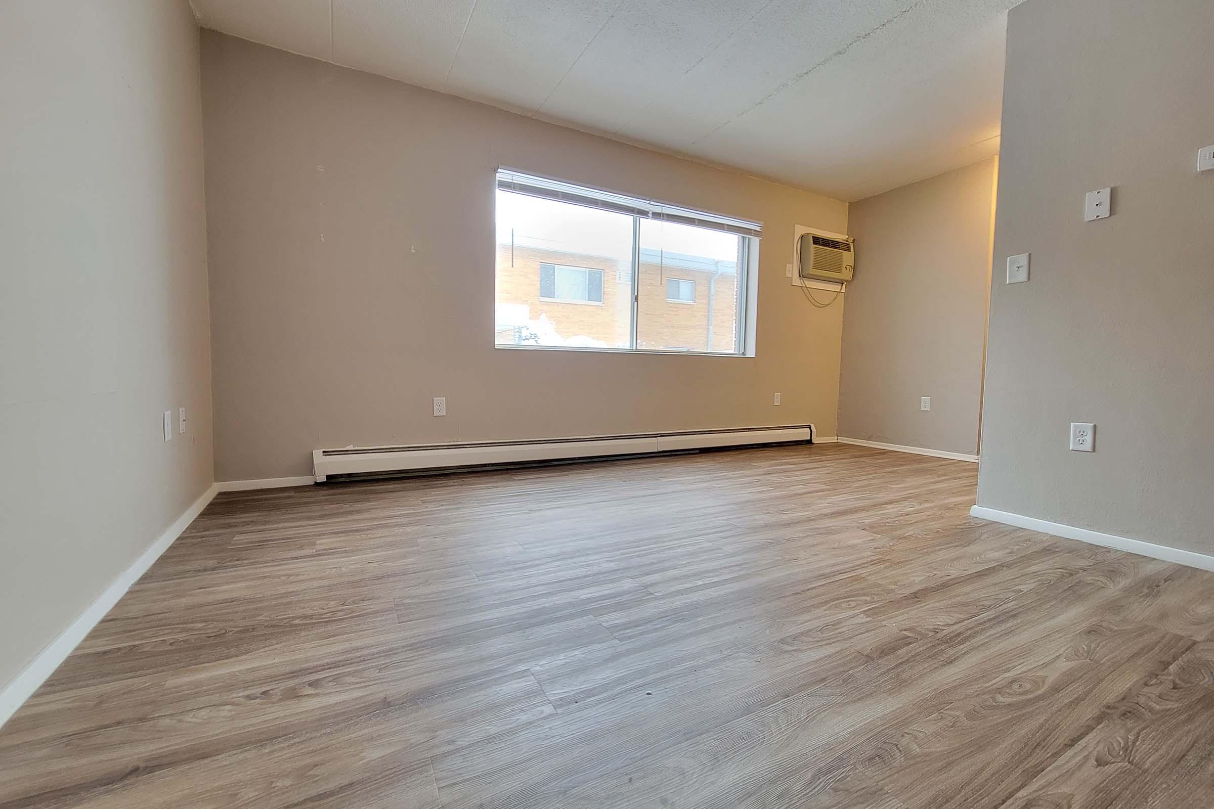 A spacious, empty room with light-colored walls and a large window letting in natural light. The floor is covered with laminate flooring, and there is an air conditioning unit on the wall. The room has a clean, modern look, ready for furniture arrangement.