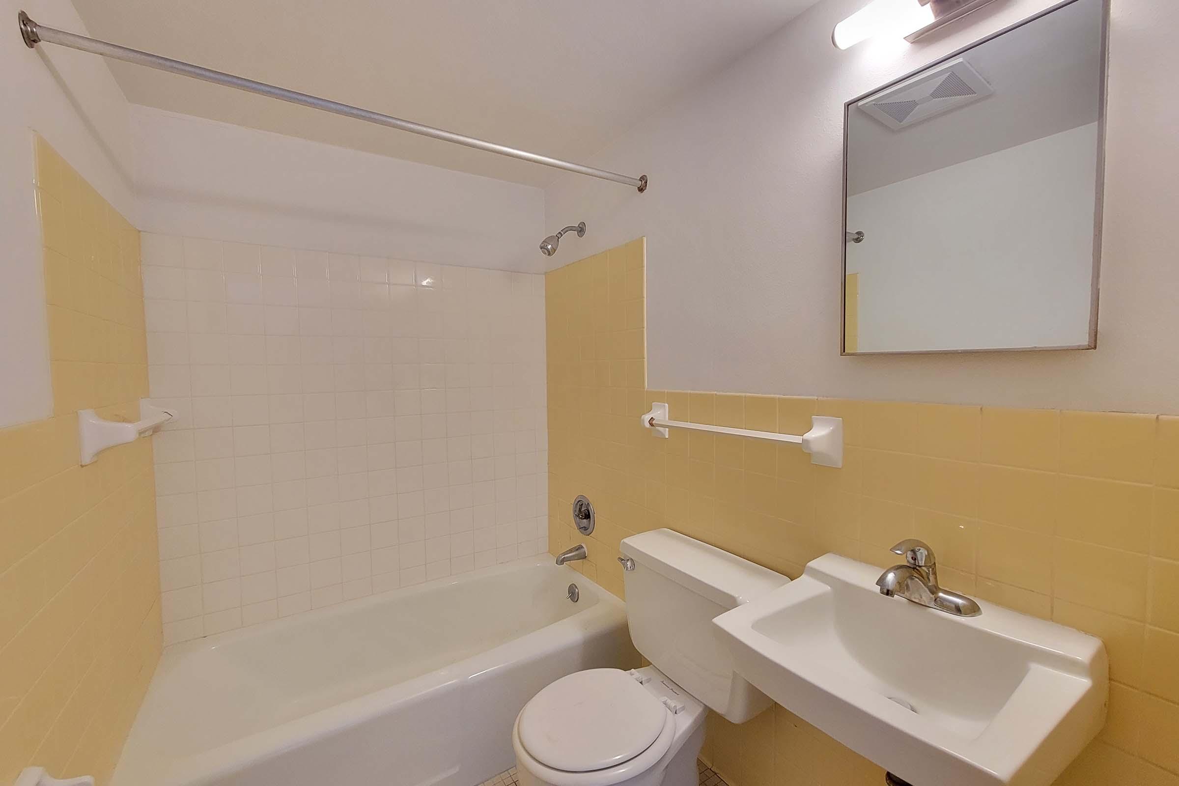 A bathroom featuring a yellow tiled bathtub with a shower, a white toilet, and a small sink mounted on the wall. There is a mirror above the sink and grab bars on the walls. The overall design is simple and includes light-colored walls and fixtures.