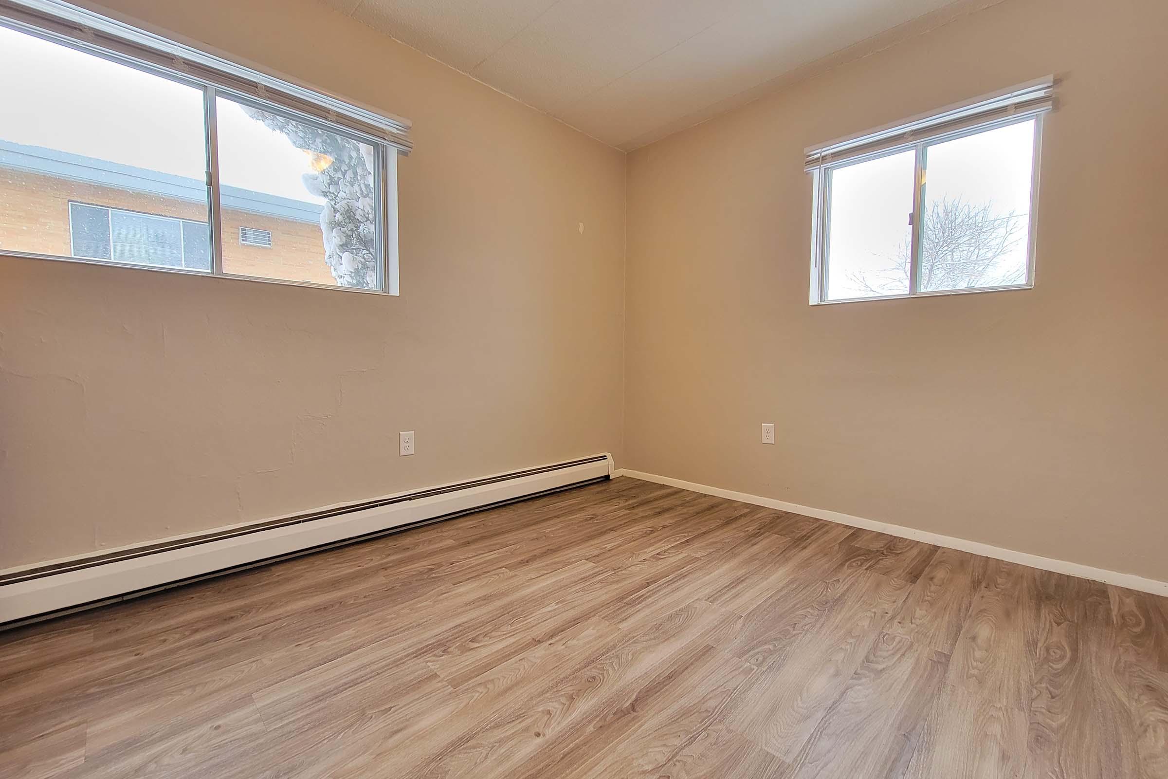 Empty room with light-colored walls and two windows. The flooring is light wood, and there are no furnishings present. One window has a view of an exterior wall, while the other reveals a clear sky. The overall space appears bright and unadorned, emphasizing the minimalistic design.